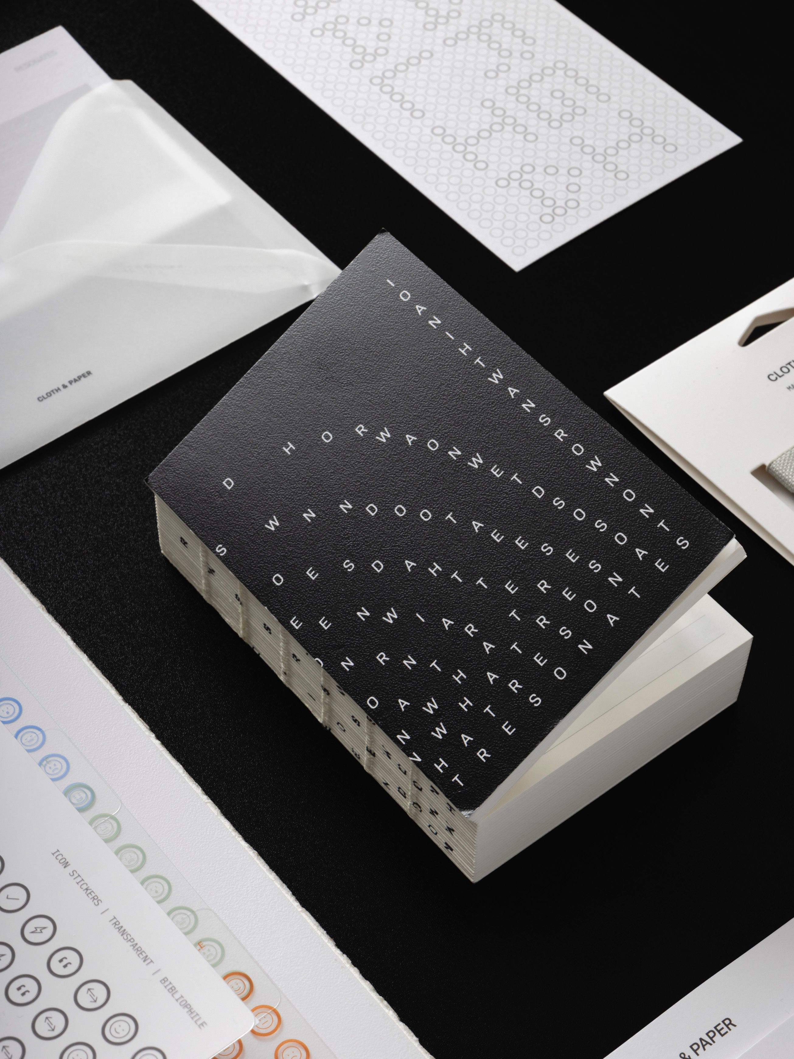 Bibliophile Notebook angled on a black surface beside a vellum envelope, a Scantron dashboard, and a packaged set of Magnetic Bookmarks.