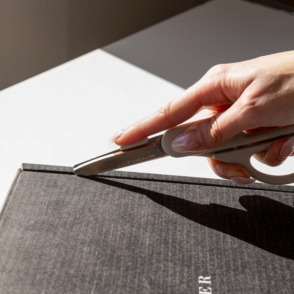 A pale hand shown using a pair of scissors in cutting mode to unseal a Cloth and Paper box.