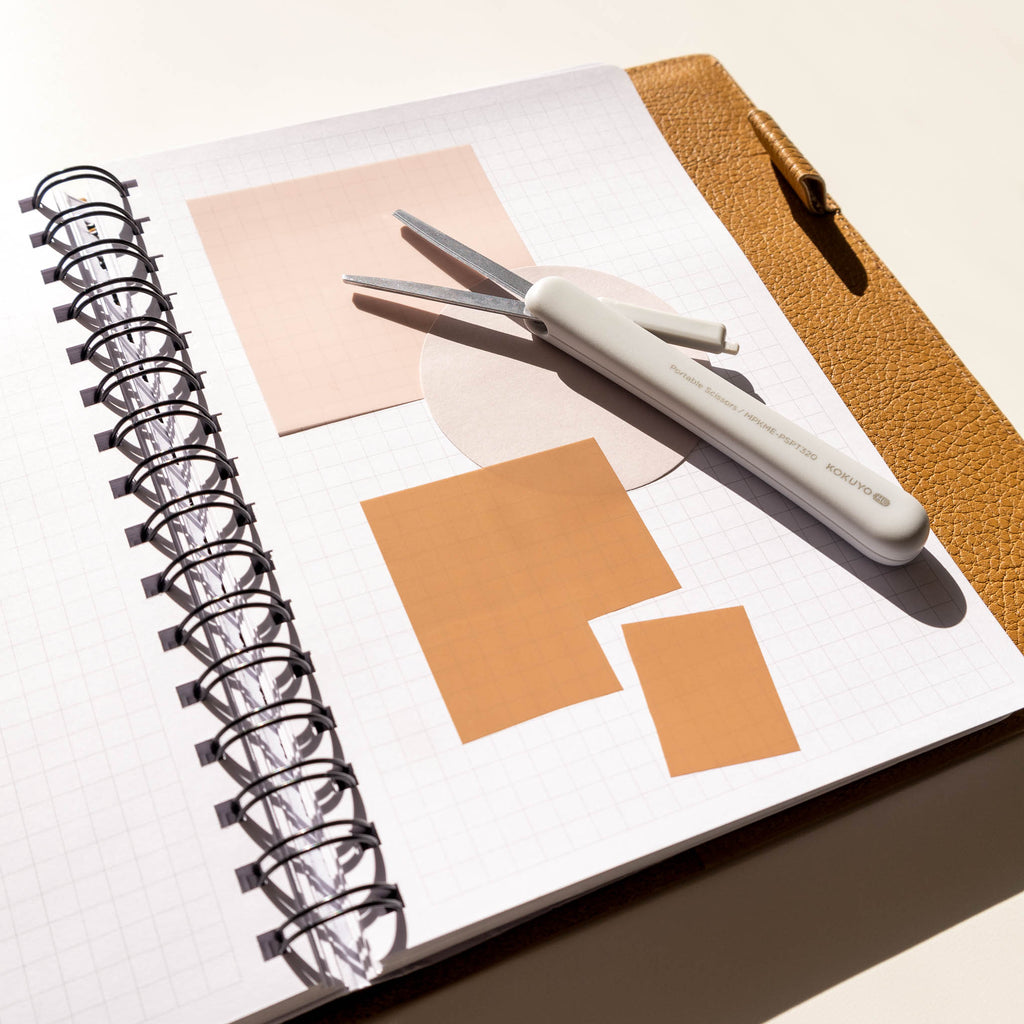 Grayish Ice scissors displayed in a notebook inside a brown leather folio. Several transparent sticky notes are layered on each other, with one being cut into a smaller square.