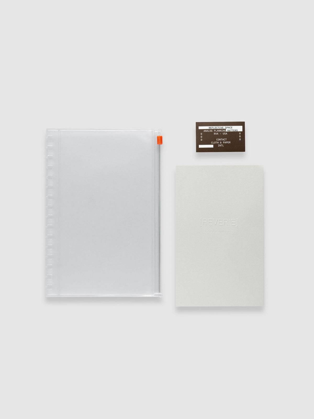 One clear zippered pouch with an orange zipper pull, one light grey embossed paper dashboard and one brown sticker laid side by side. Shown in size A5 Spiral.