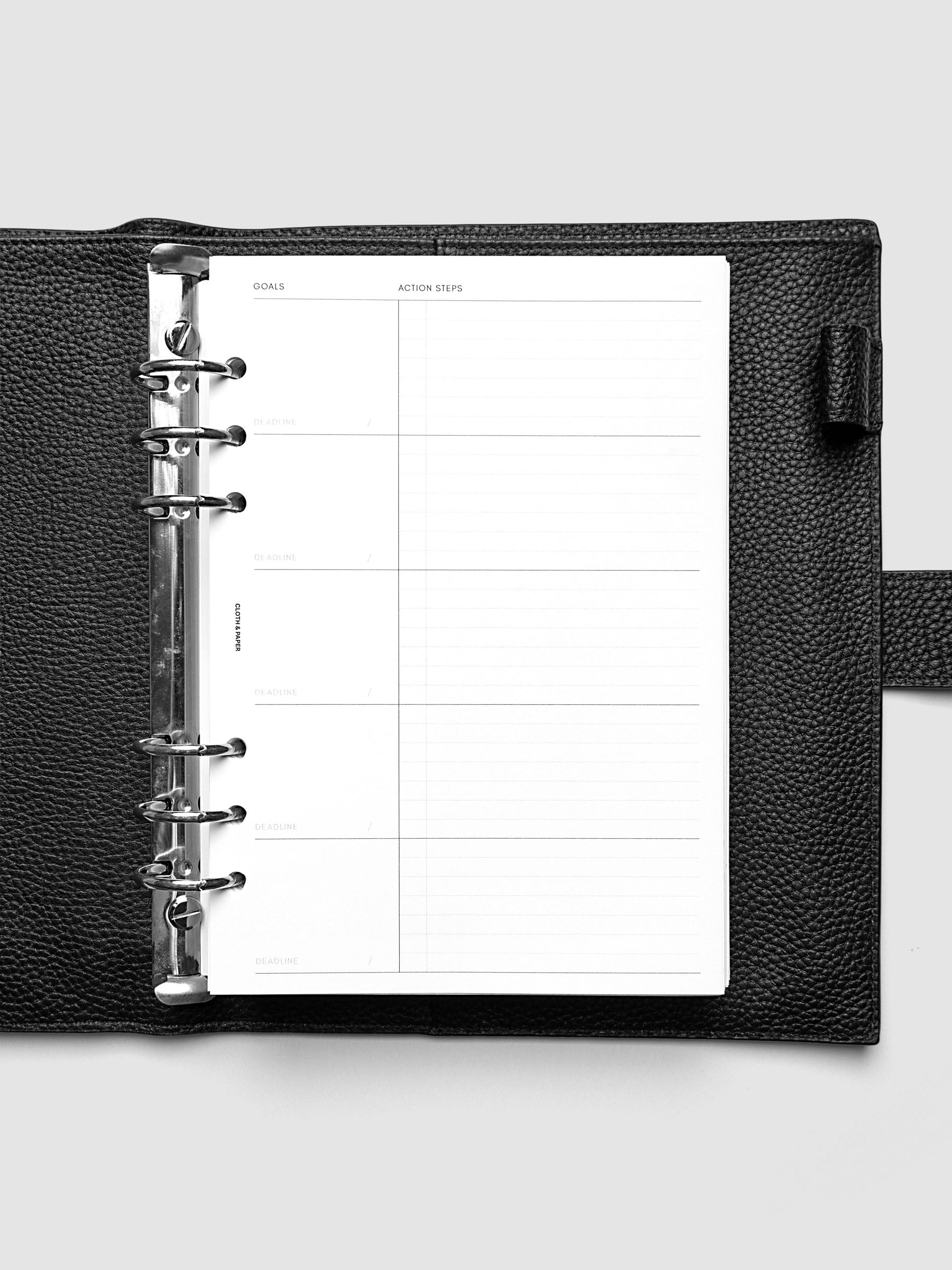 Open black leather planner displaying a blank goals and action steps worksheet with lined sections. The planner is laid flat on a light gray surface.