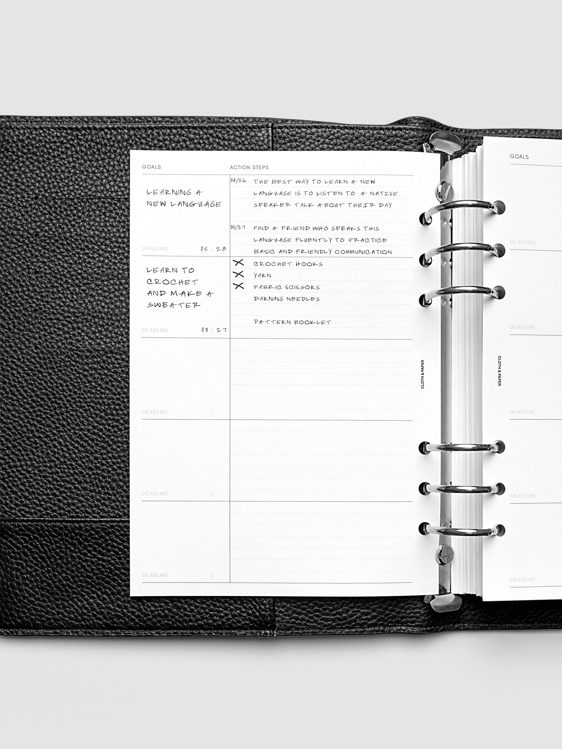 Open black leather planner displaying a goals and action steps page, with handwritten notes about learning a new language and blogging. The planner is open on a white background, showing organized sections and a metal ring binder.