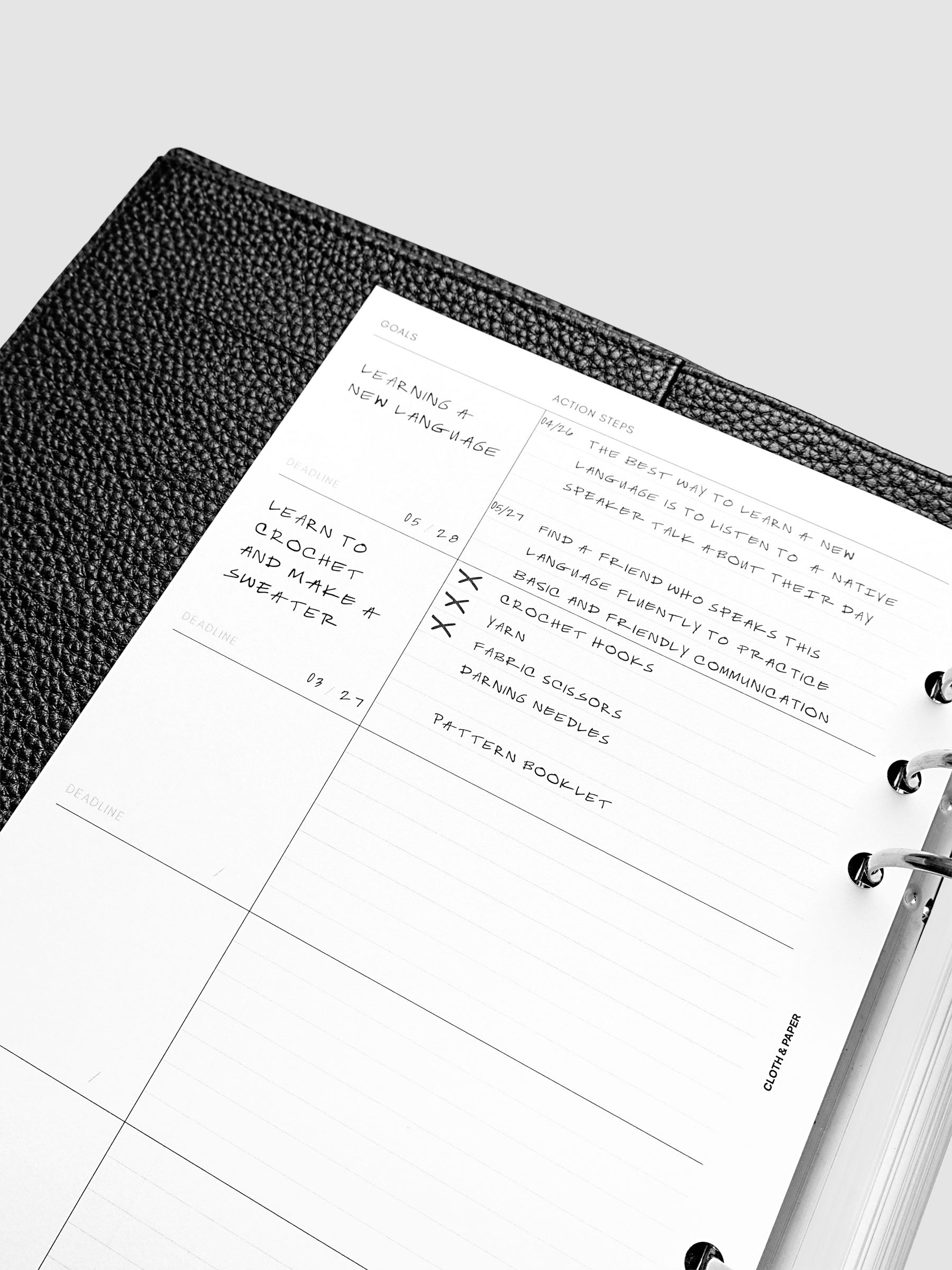 Open planner showing handwritten goals and action steps for learning a new language, including tasks like finding a study partner and using workbooks. The planner rests on a textured black surface.