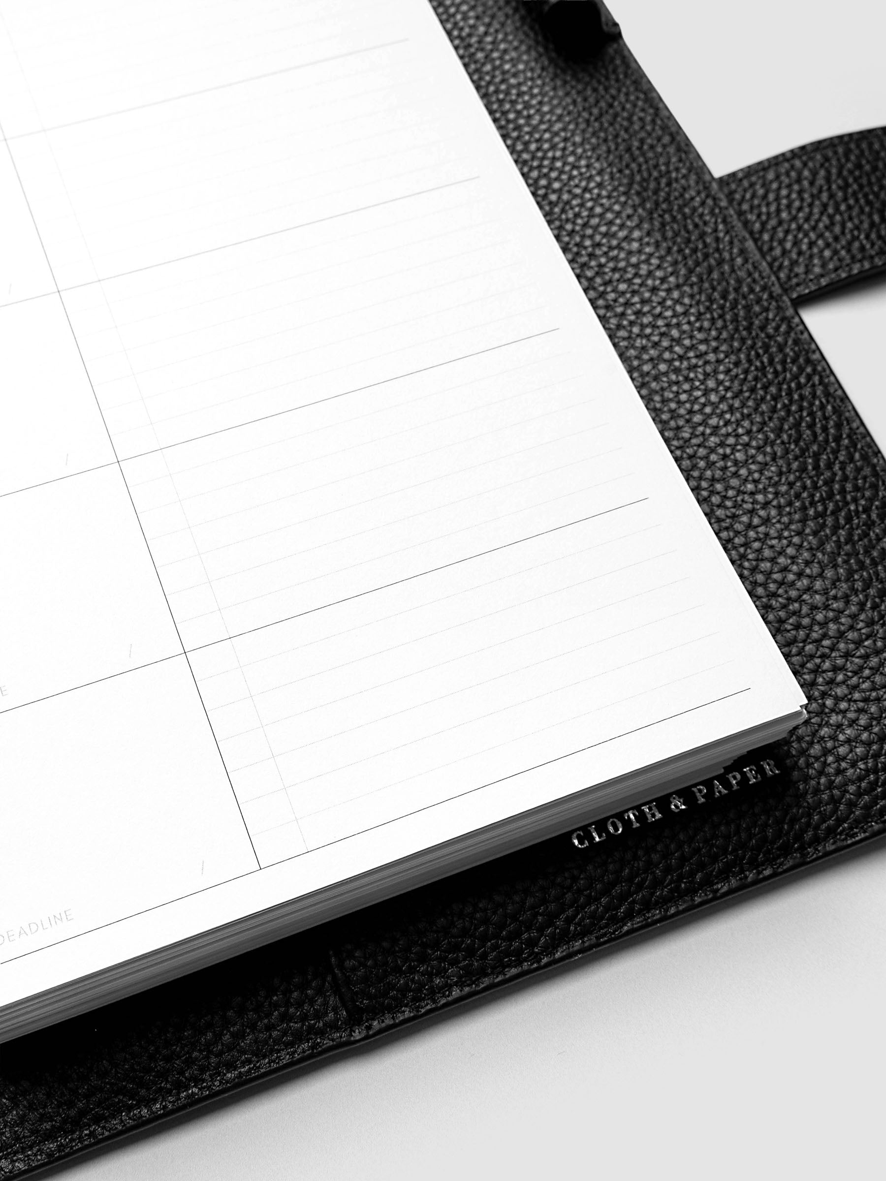 Close-up of an open planner with lined pages resting on a textured black leather cover. The words "CLOTH & PAPER" are embossed on the cover's edge.