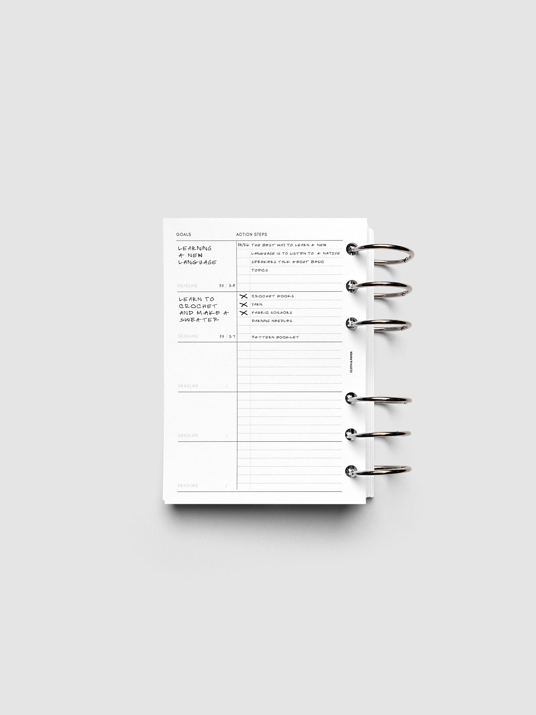 Open ring-bound planner displaying a goals and action steps page with sections for cleaning, language, and tactical project tasks. The planner is placed on a plain light gray background.