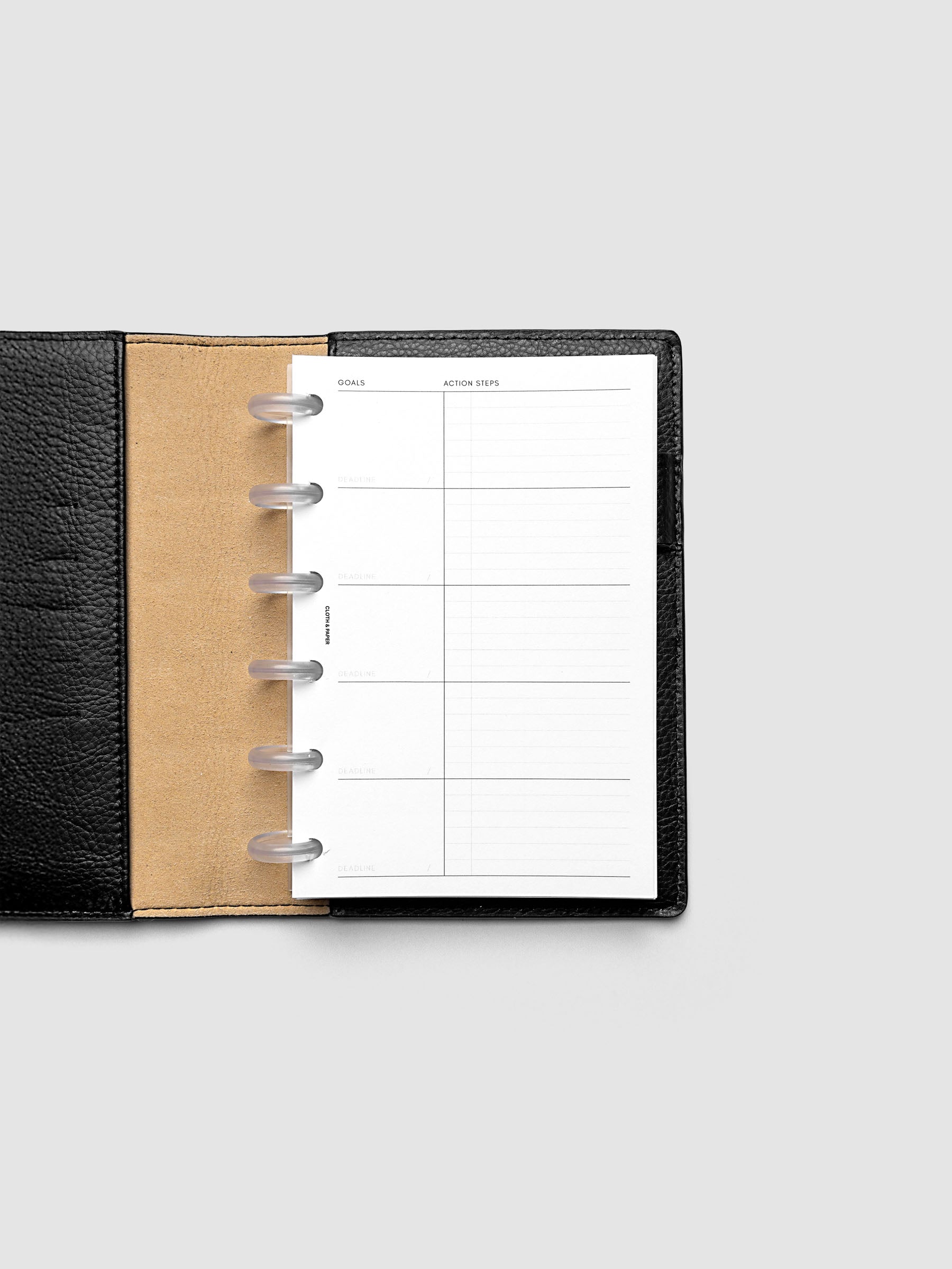 Open black leather planner displaying a blank weekly goals and action steps page. The planner is set against a plain light gray background.