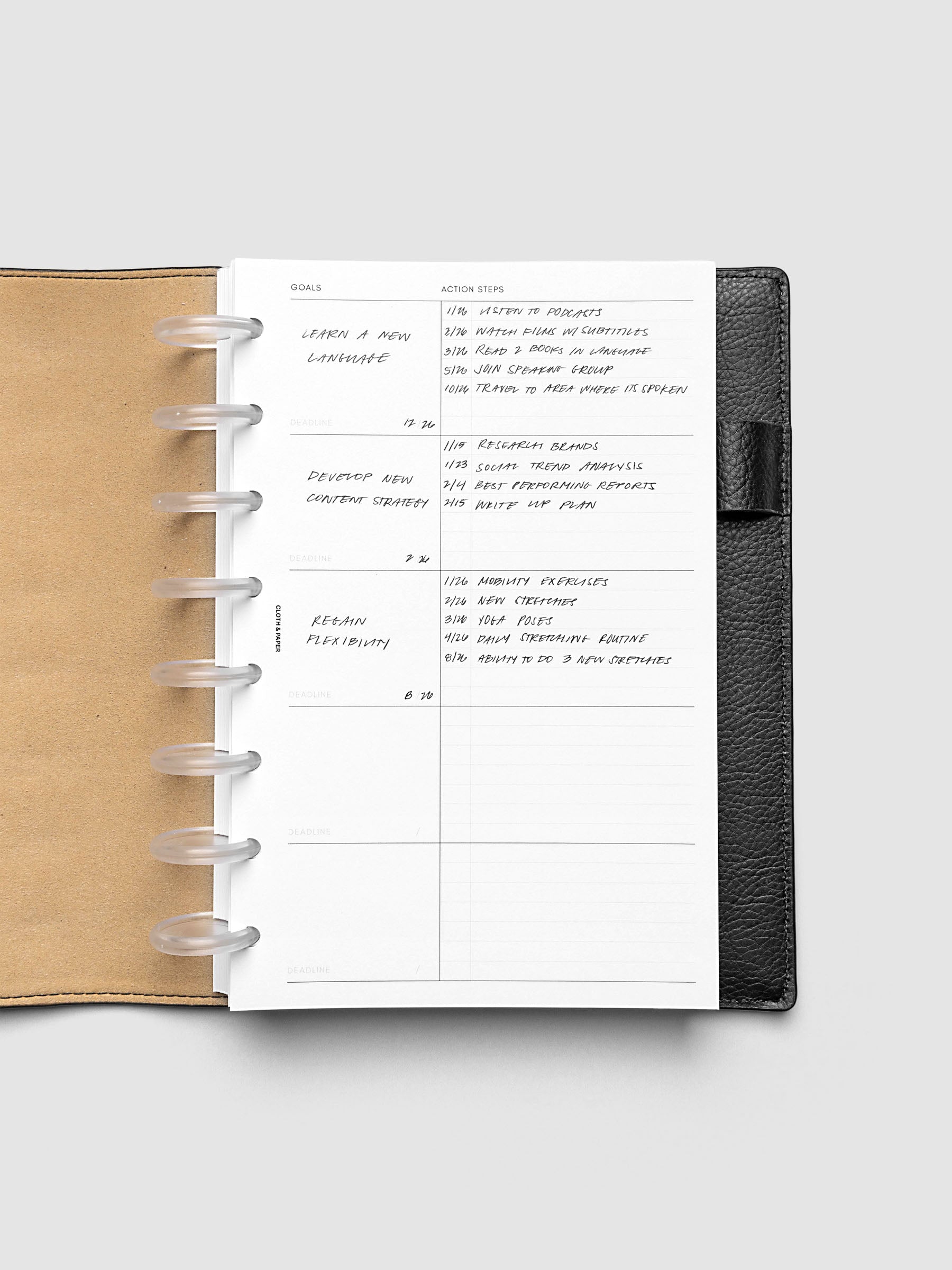 Open leather-bound planner displaying a handwritten goals and action list page with tasks organized by date. The planner is laid flat on a light gray background.