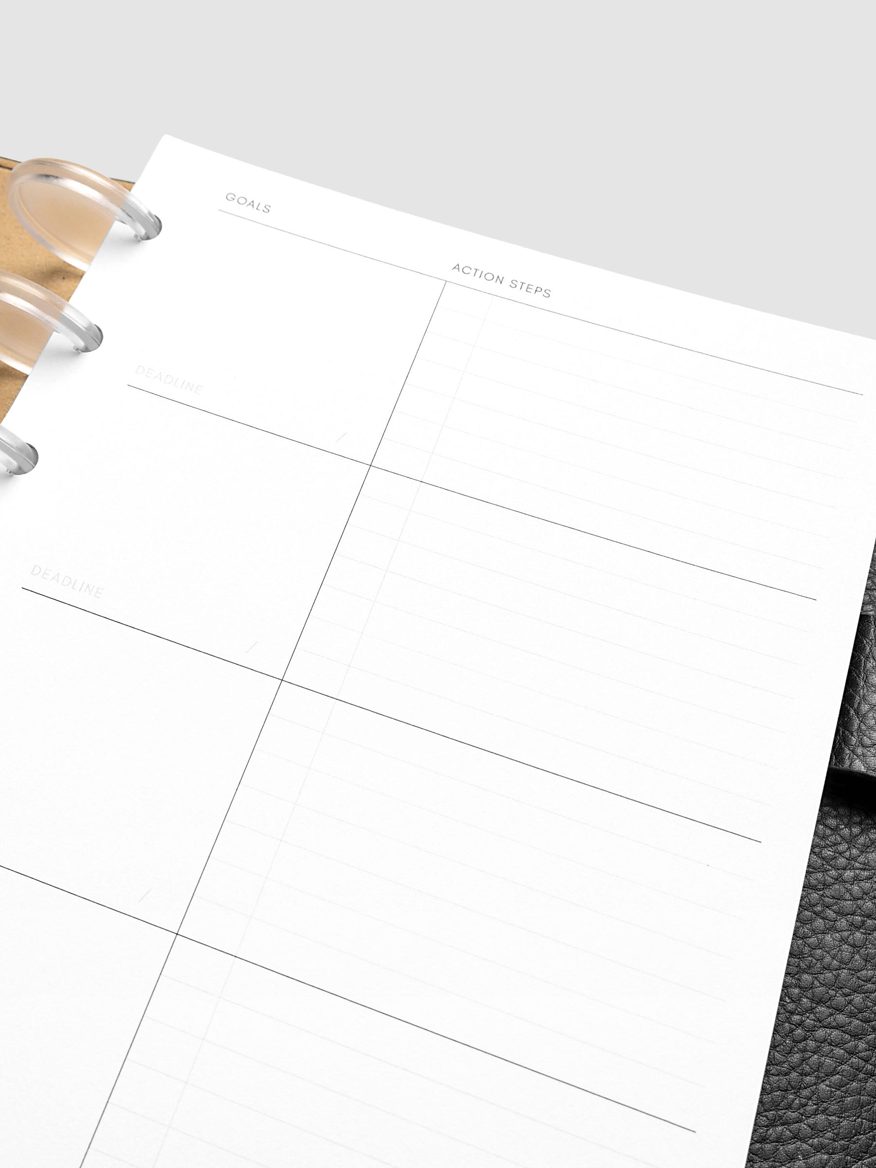 Close-up of a blank planner page with sections labeled "Goals," "Action Steps," and "Deadline." The planner rests on a gray leather surface.