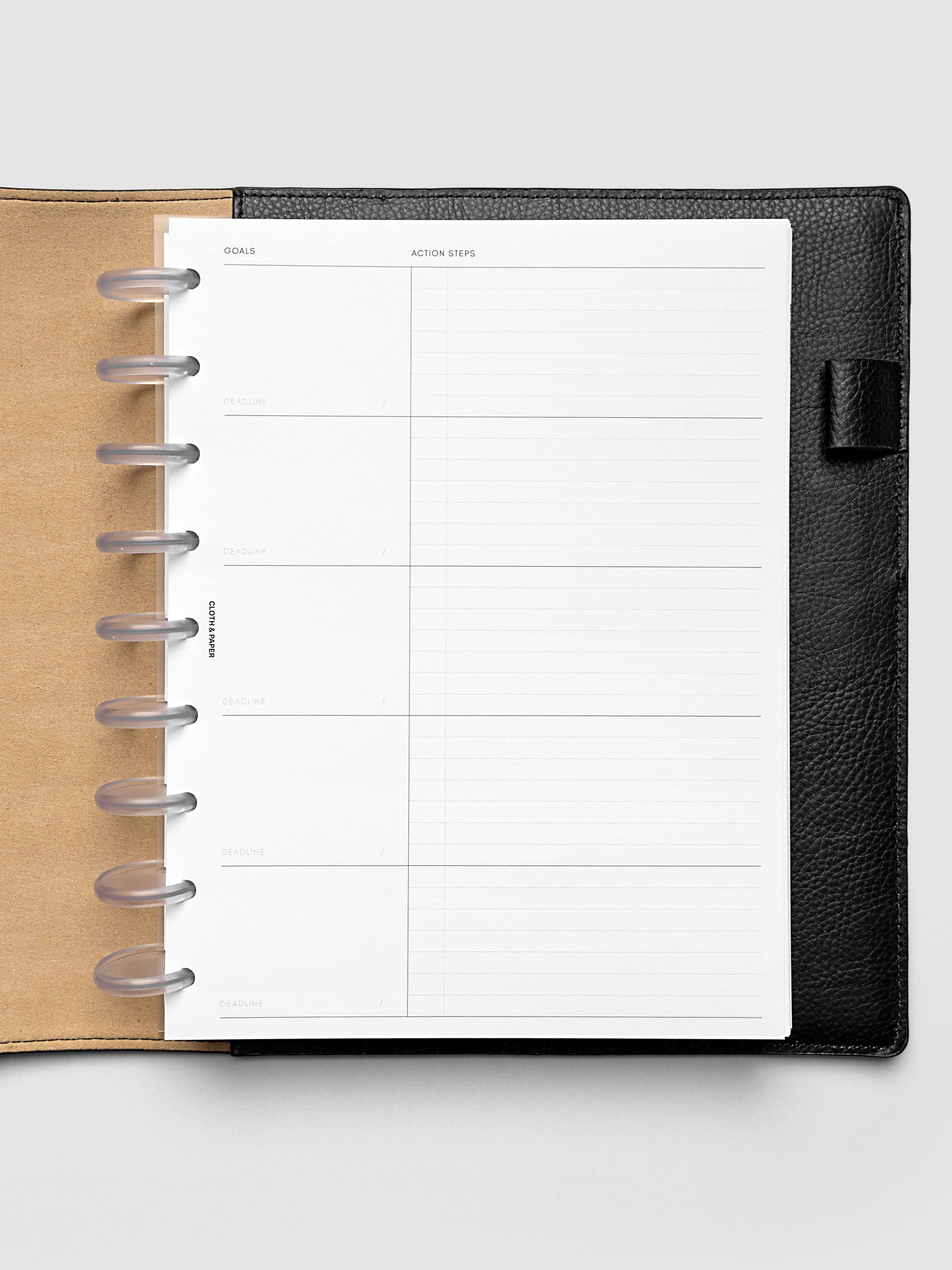Open black leather planner displaying a blank goals and action steps page with lined and sectioned areas for writing. The planner is laid flat on a light gray background.