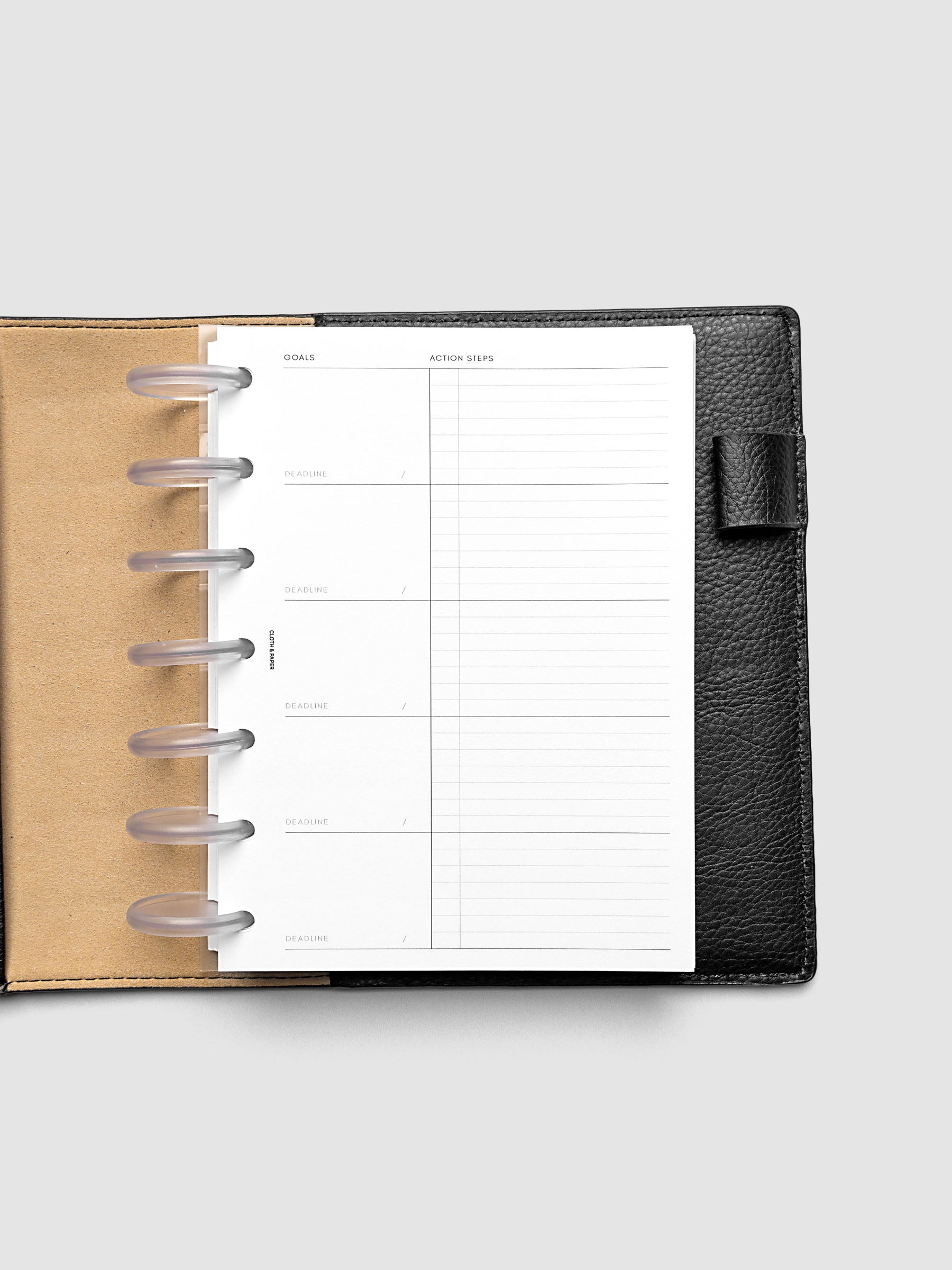 Open black leather planner displaying a blank goals and action steps page with ring binding. The planner is laid flat on a light gray background.