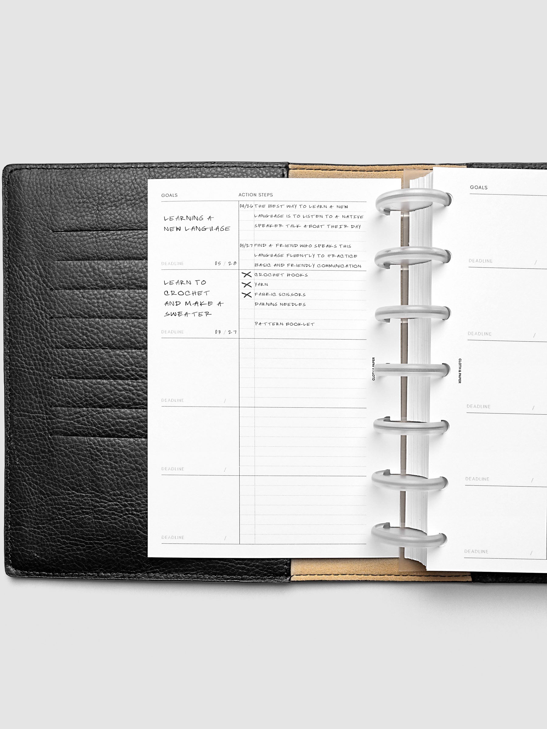 Open black leather planner displaying a goals and action items page, with handwritten notes and checkmarks on the left side. The right page is blank with printed sections for goals and deadlines.