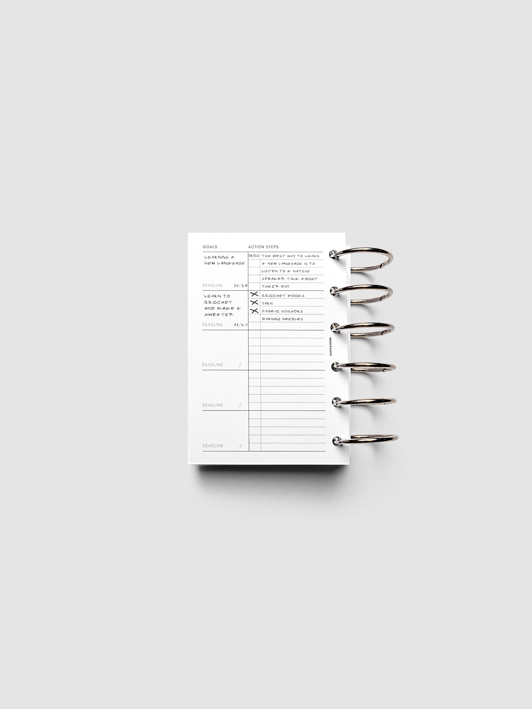 Open ring-bound planner with sections labeled "Goals" and "Action Steps" on a white background. The planner has sample text filled in.