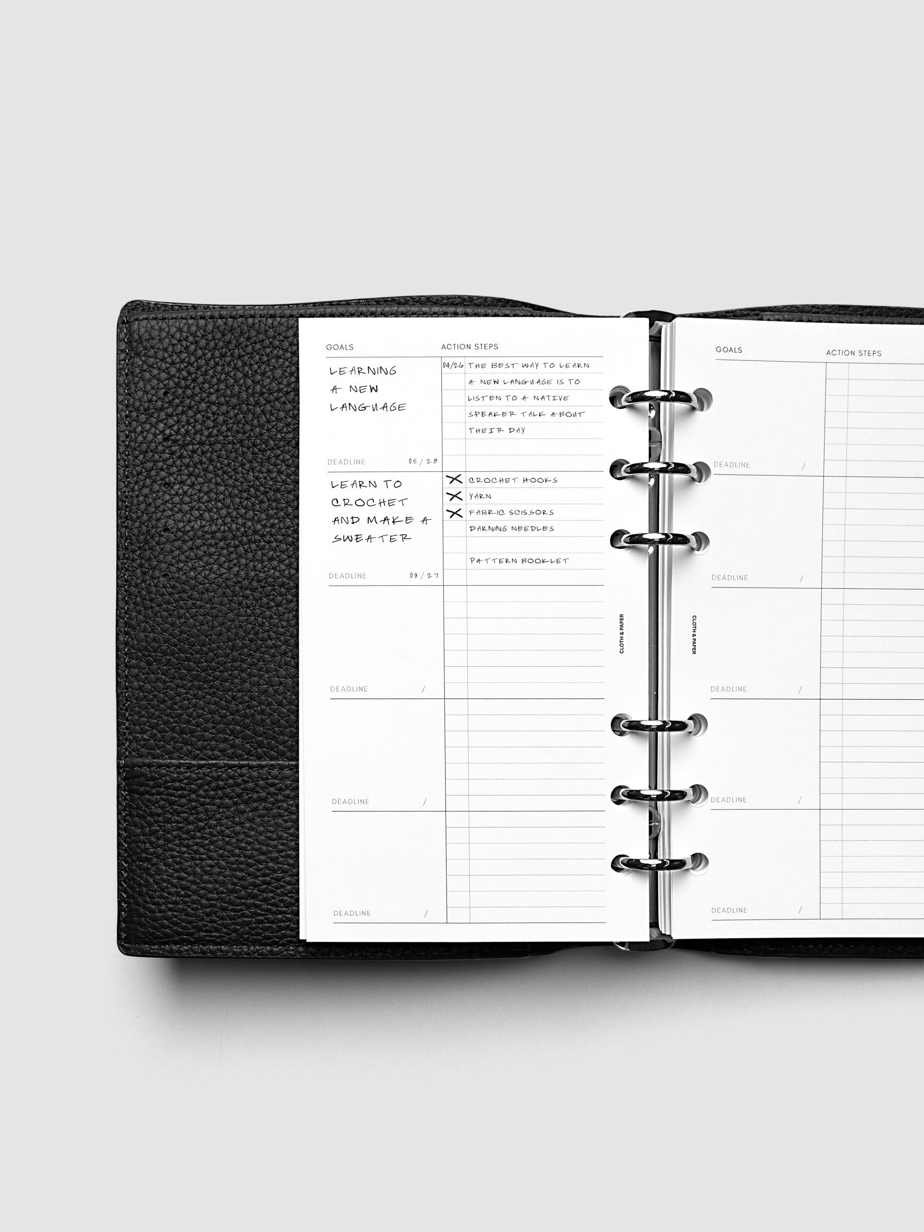 Open black leather planner displaying a goals and action steps page, with handwritten notes about learning a new language and tracking progress. The planner is laid flat on a light gray surface.