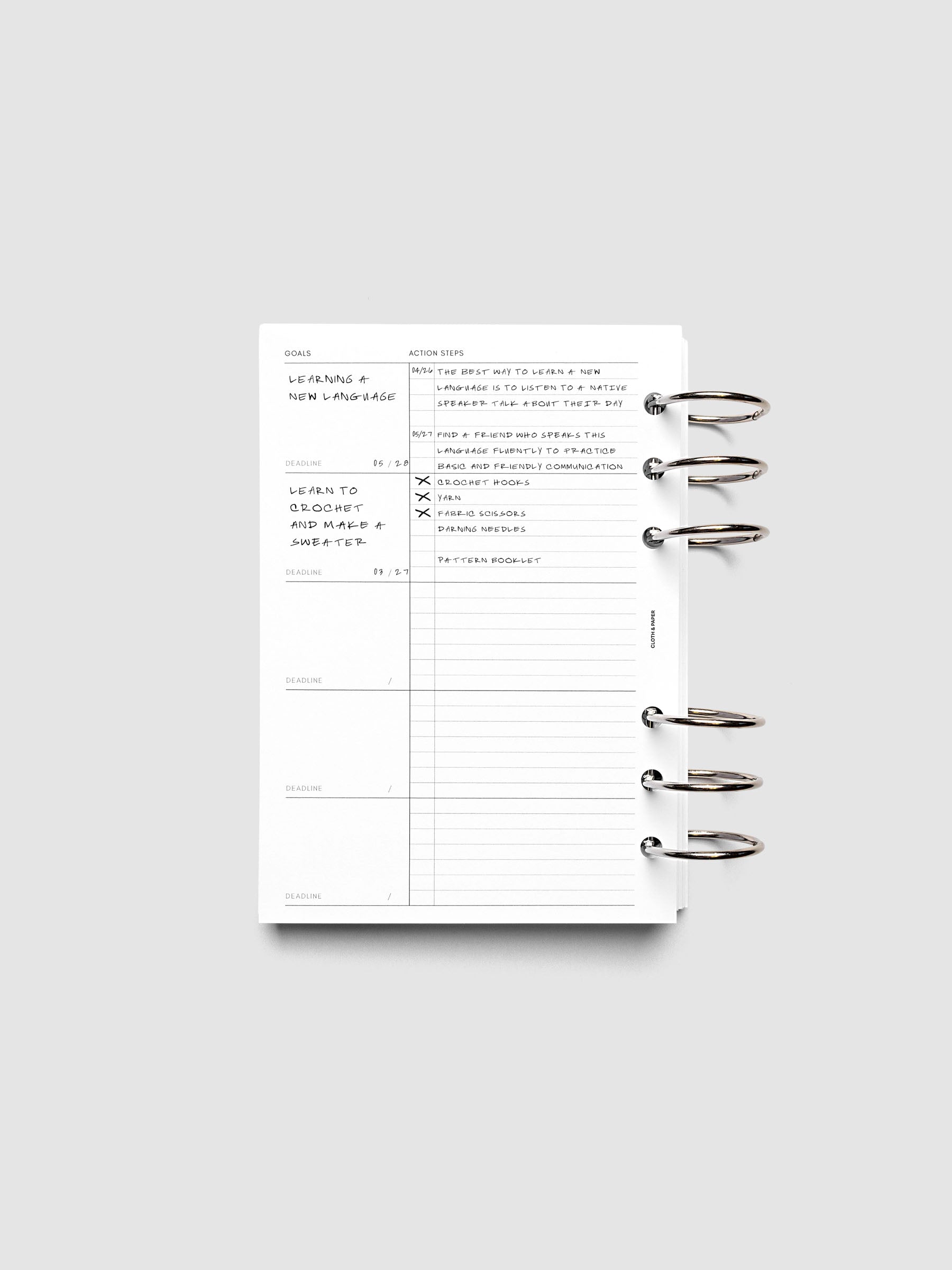 Open ring-bound planner displaying a goals and action list page, with handwritten notes about learning a new language and steps to achieve it. The planner is laid flat on a light gray background.