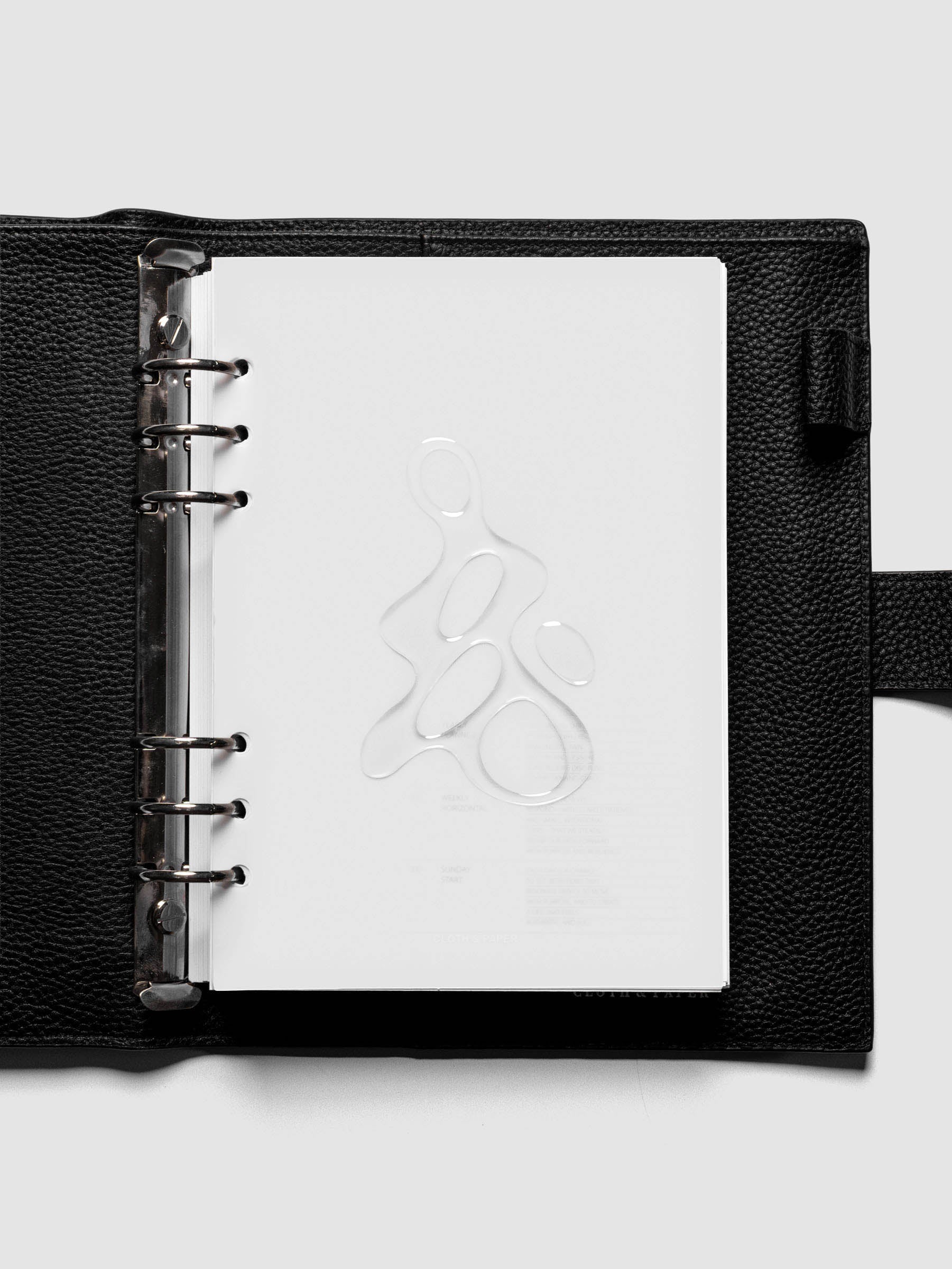 Light grey, semi-transparent, and glossy plastic planner dashboard with organic raised shape design in size A5 shown ringbound in a black leather agenda.