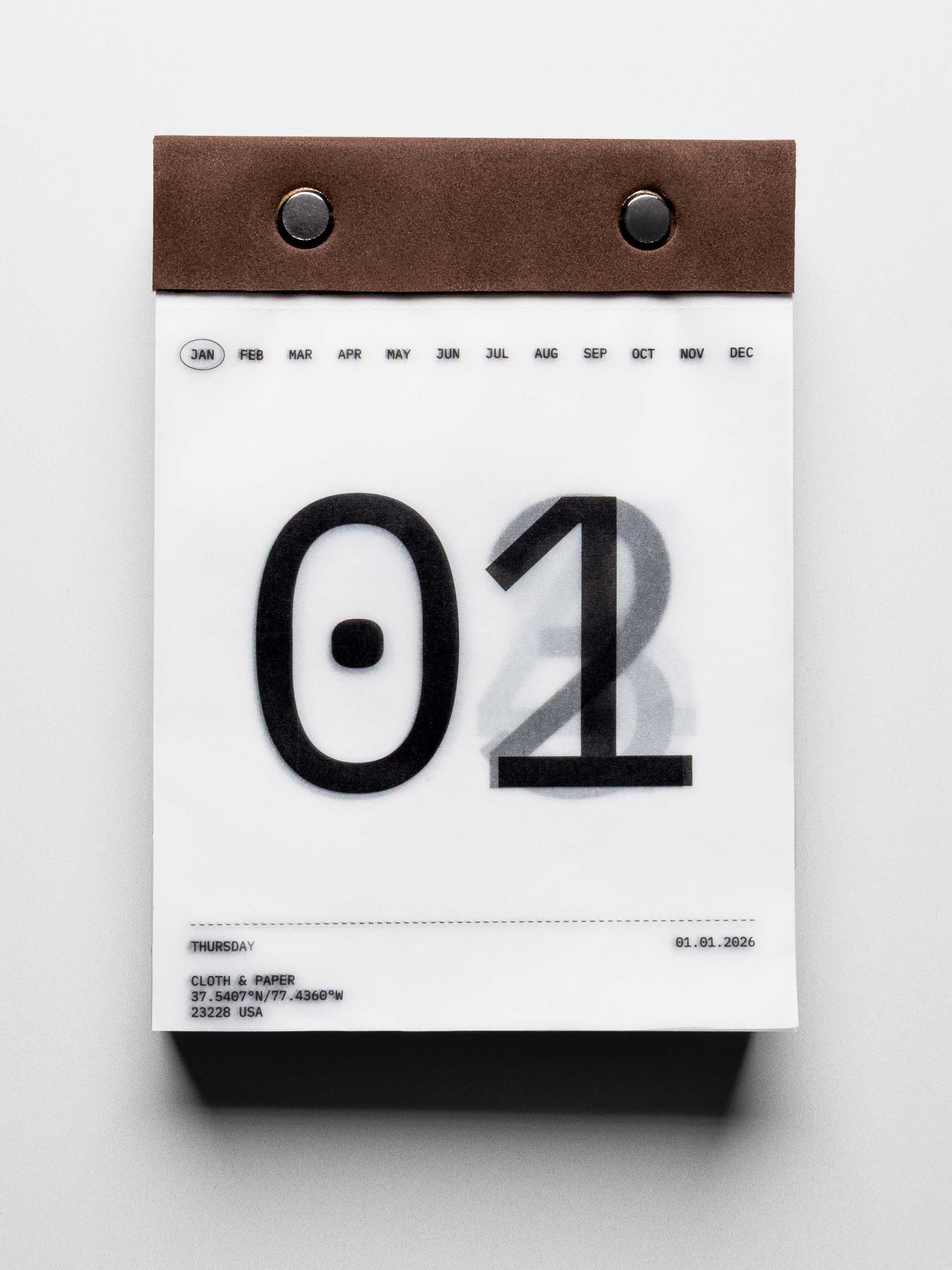 Vellum paper calendar tear-off desk pad bound with suede and metal pins. Page shows January 1st in large typographic design with days below slightly visible through transparent paper.