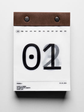 Vellum paper calendar tear-off desk pad bound with suede and metal pins. Page shows January 1st in large typographic design with days below slightly visible through transparent paper.