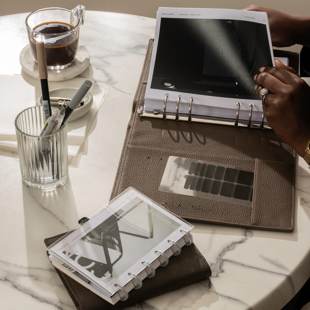 A left hand is flipping through a set of 2024 planner inserts that are inside a leather planner.  There is a cup of coffee on the table and other surrounding planner accessories.