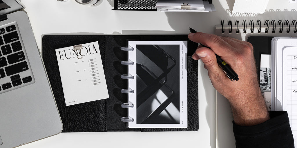 A right hand is holding a pen next to a leather planner filled with 2024 planner inserts. There is an edge of a laptop computer on the left. 