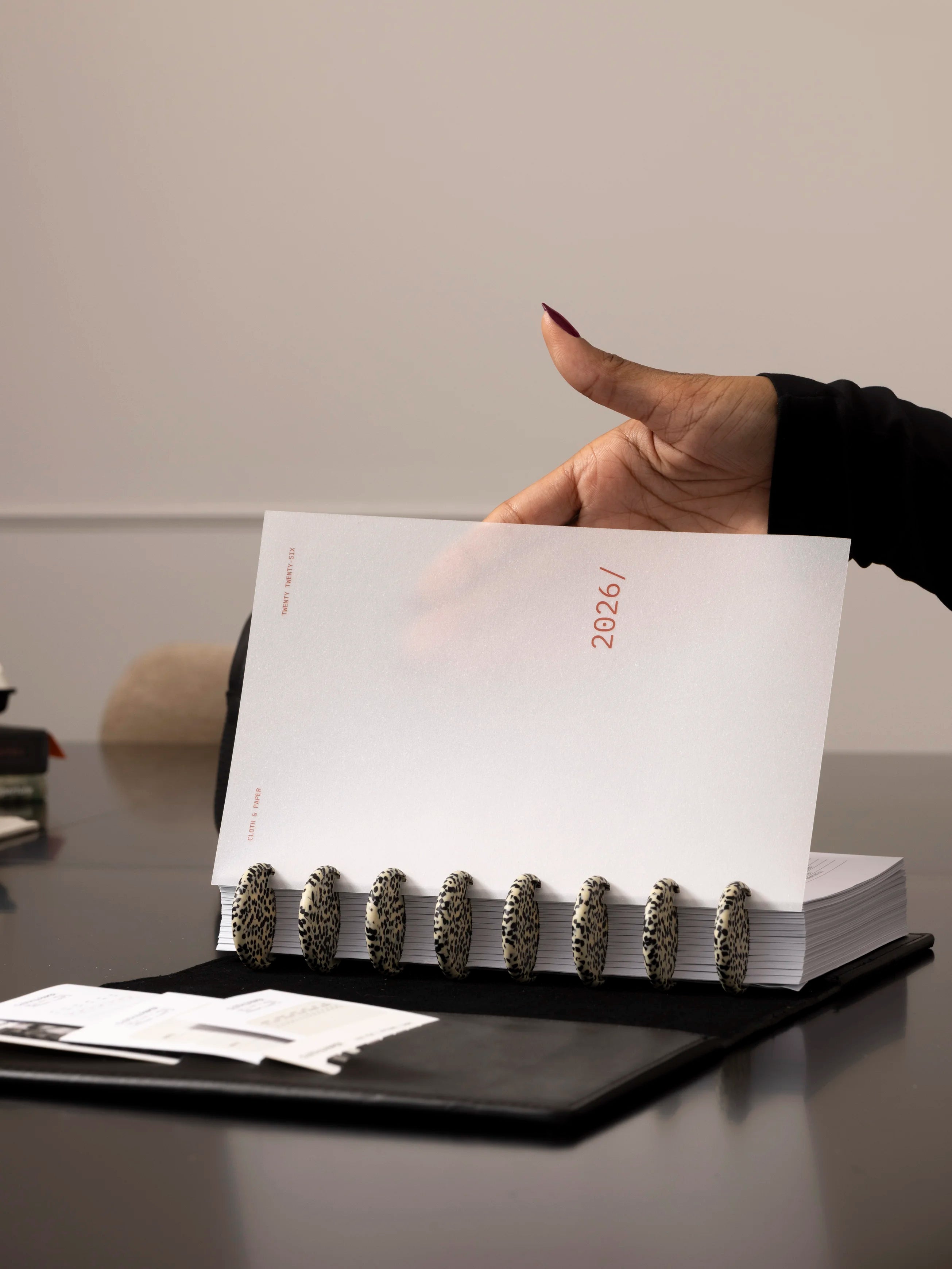 Hand flipping through a planner with bold speckled discs and a 2026 cover page inside a black leather folio.