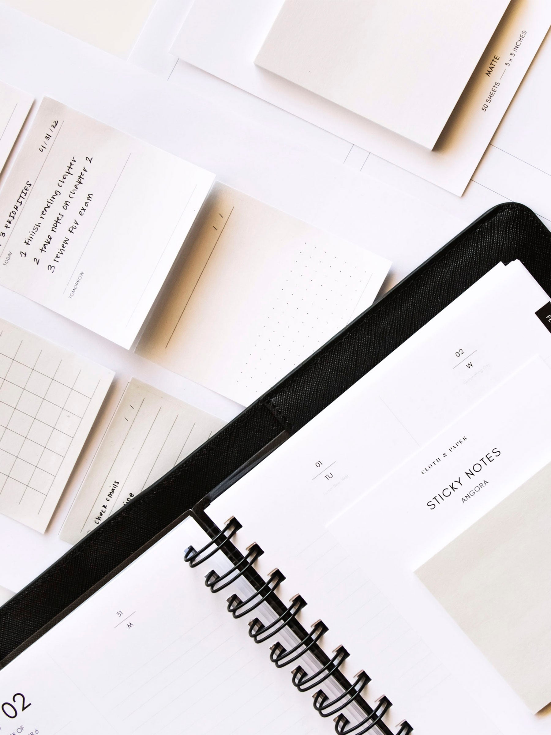 Pad of Note Neutrality Sticky Notes resting on spiral bound planner inside leather cover. A Uni Pin is in the cover's pen loop, and printed sticky notes and another pad of blank sticky notes rest on the table underneath the planner.