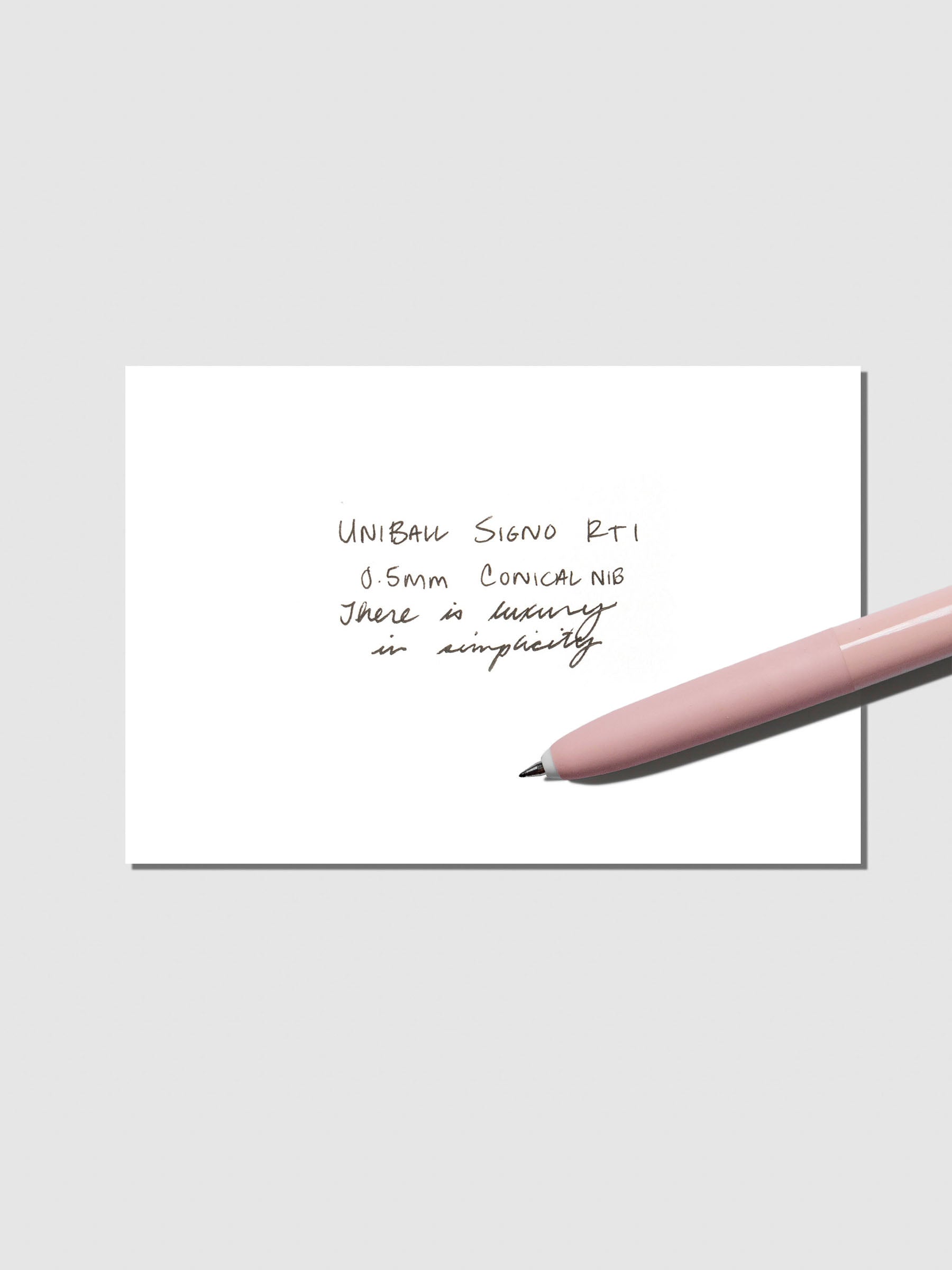 Uni Ball Signo in Shell Pink displayed with writing sample on a neutral background.