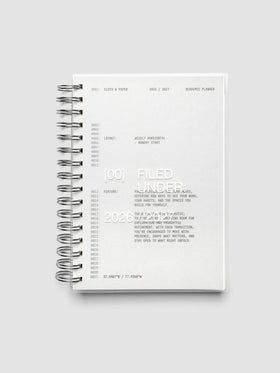 Transparent spiral-bound academic planner closed, showing the grey cardstock dashboard through the clear plastic cover, with silver spiral binding along the left edge
