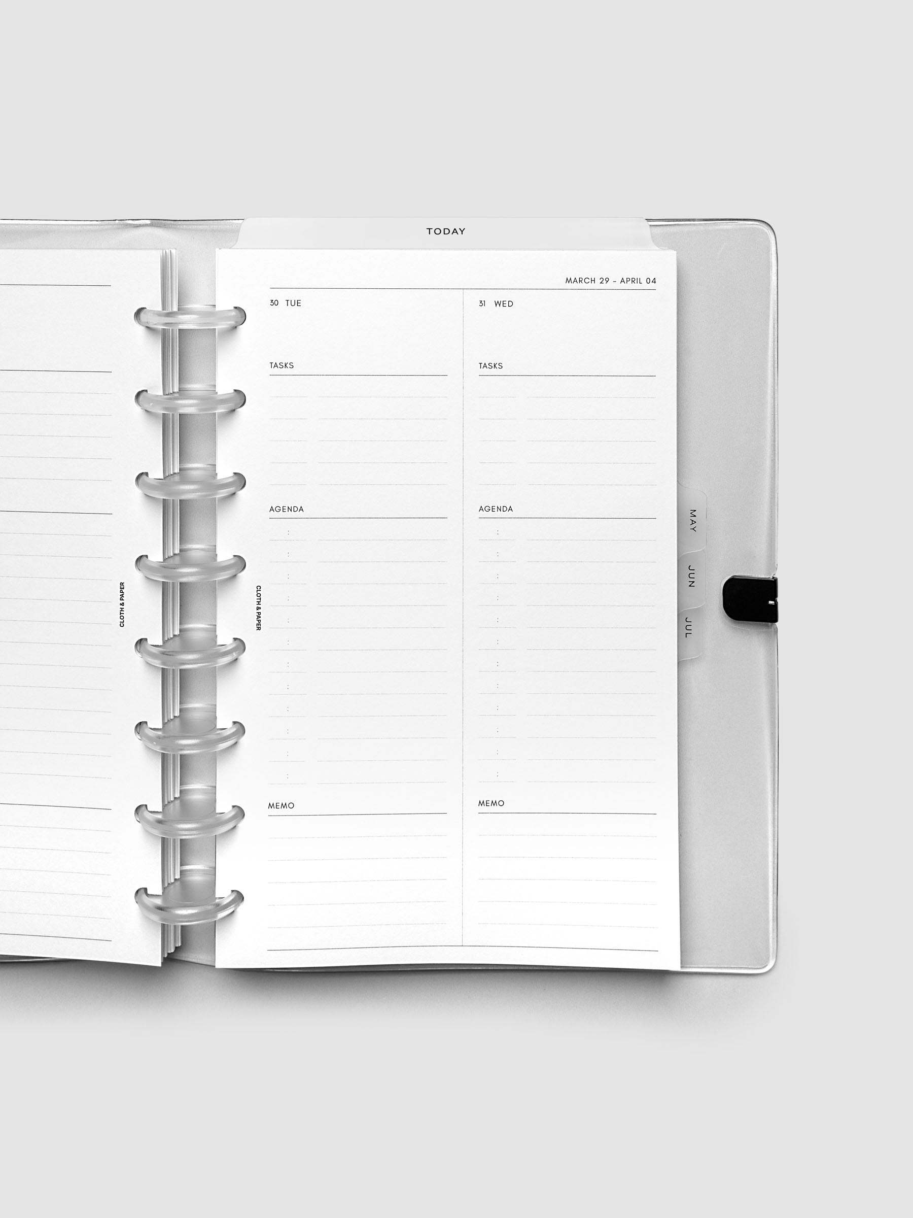 Close-up of an open planner with clear 8-disc binding, weekly spread showing Tuesday and Wednesday daily columns with tasks, agenda, and memo sections, tabbed monthly dividers visible at the edge, Half Letter 8-Disc Punch size