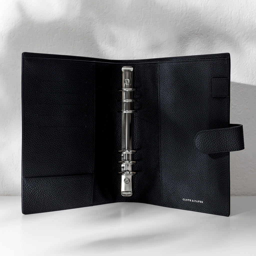Black leather agenda displayed on an off-white background. It is opened to show off the silver hardware and company logo stamp.