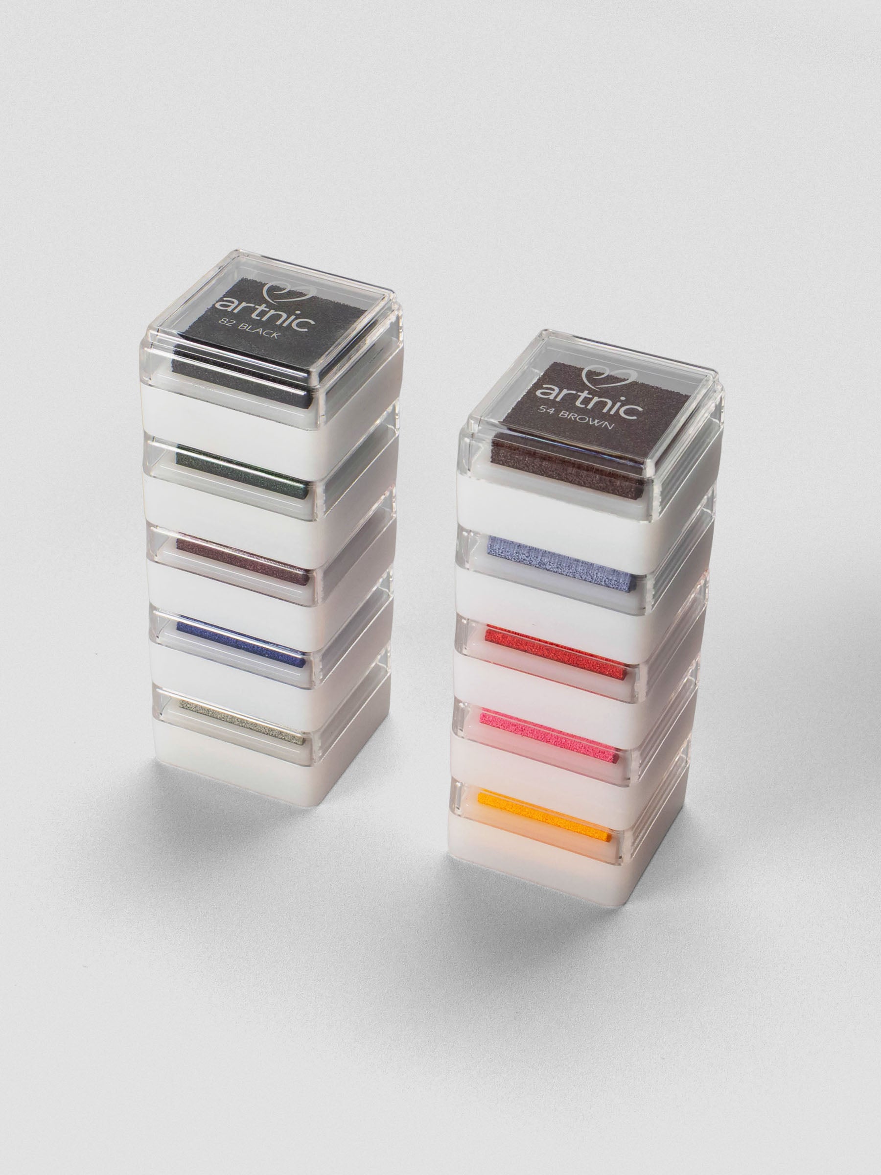 Two stacks of Artnic Stamp Ink Pads in assorted colors, each topped with a labeled clear lid showing the color name, displayed on a light background.