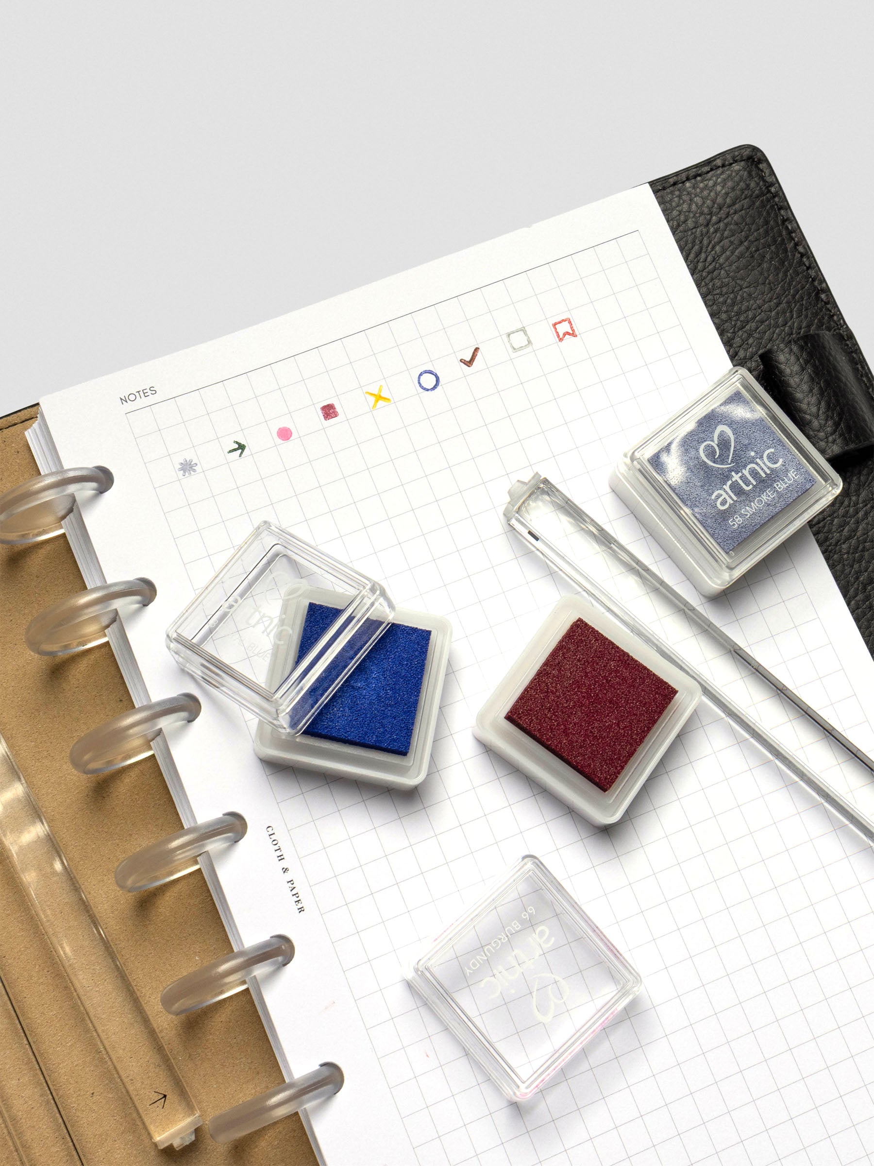 Open Artnic Stamp Ink Pads in blue and burgundy resting on a planner page with stamped icons, clear lids placed nearby, and a Stamp Stick visible.