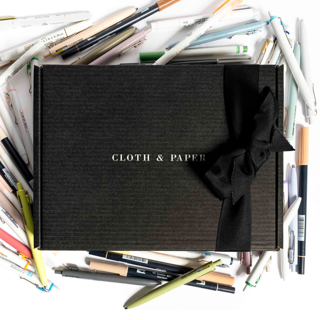 Cloth and Paper box adorned with a black bow displayed on a white background. A large collection of pens are arranged around the box.