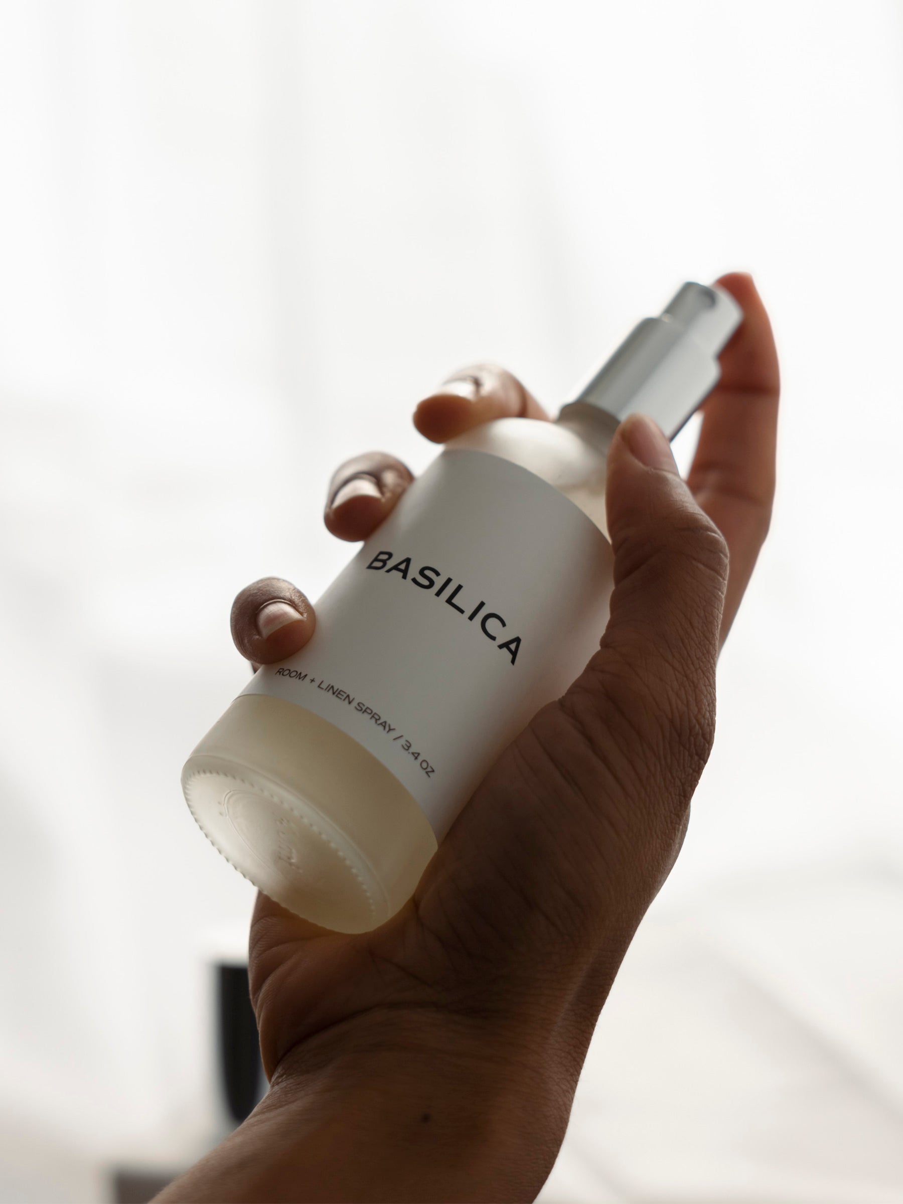 Spray shown uncapped, held in a hand about to push down the spray mechanism. The lighting is soft and cast from the back.