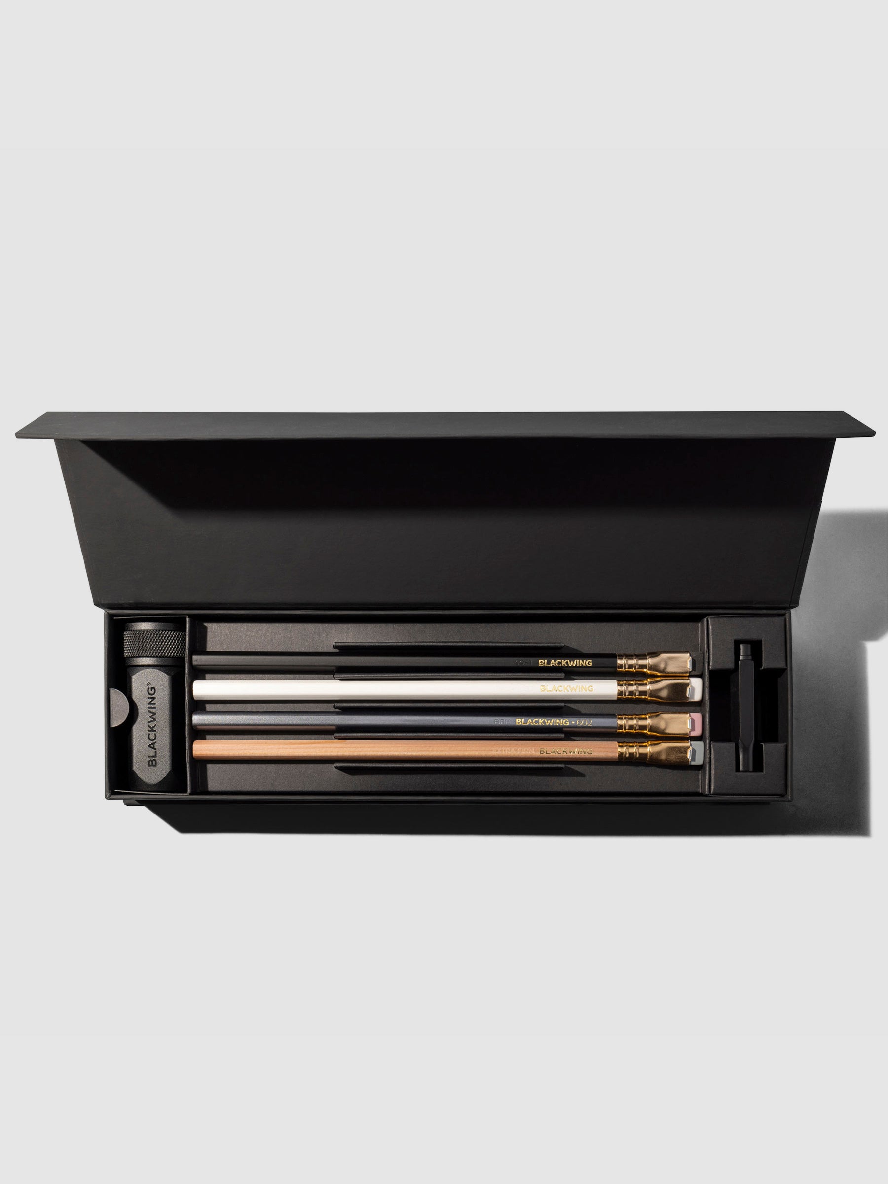 Box displayed on a neutral background. It is open, showcasing four pencils, a black point guard, and black pencil sharpener.