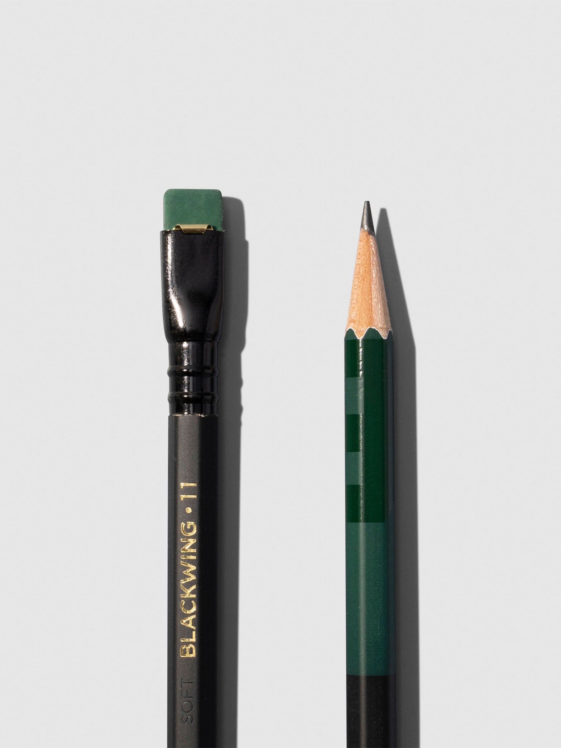 Closeup of two black and green wooden pencils displayed on a neutral background. One is shown eraser side up, the other point side up.