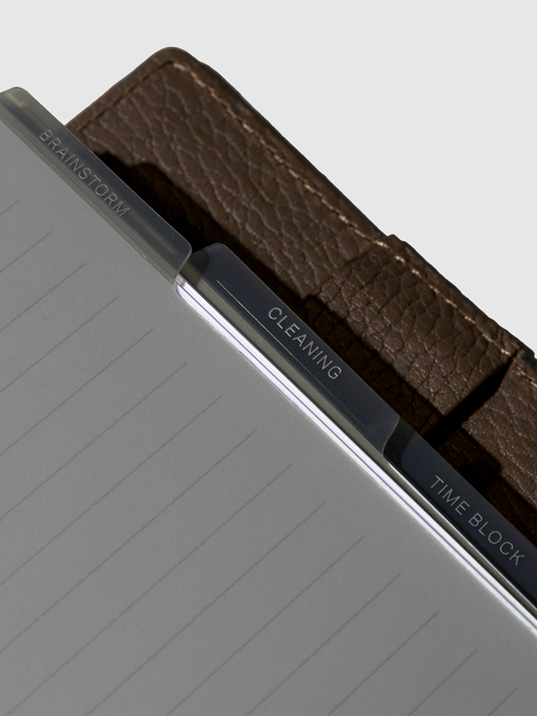 A close up image of the tinted gray dividers in use inside a brown leather planner. The divider tabs have labels on them that say brainstorm, cleaning and time block