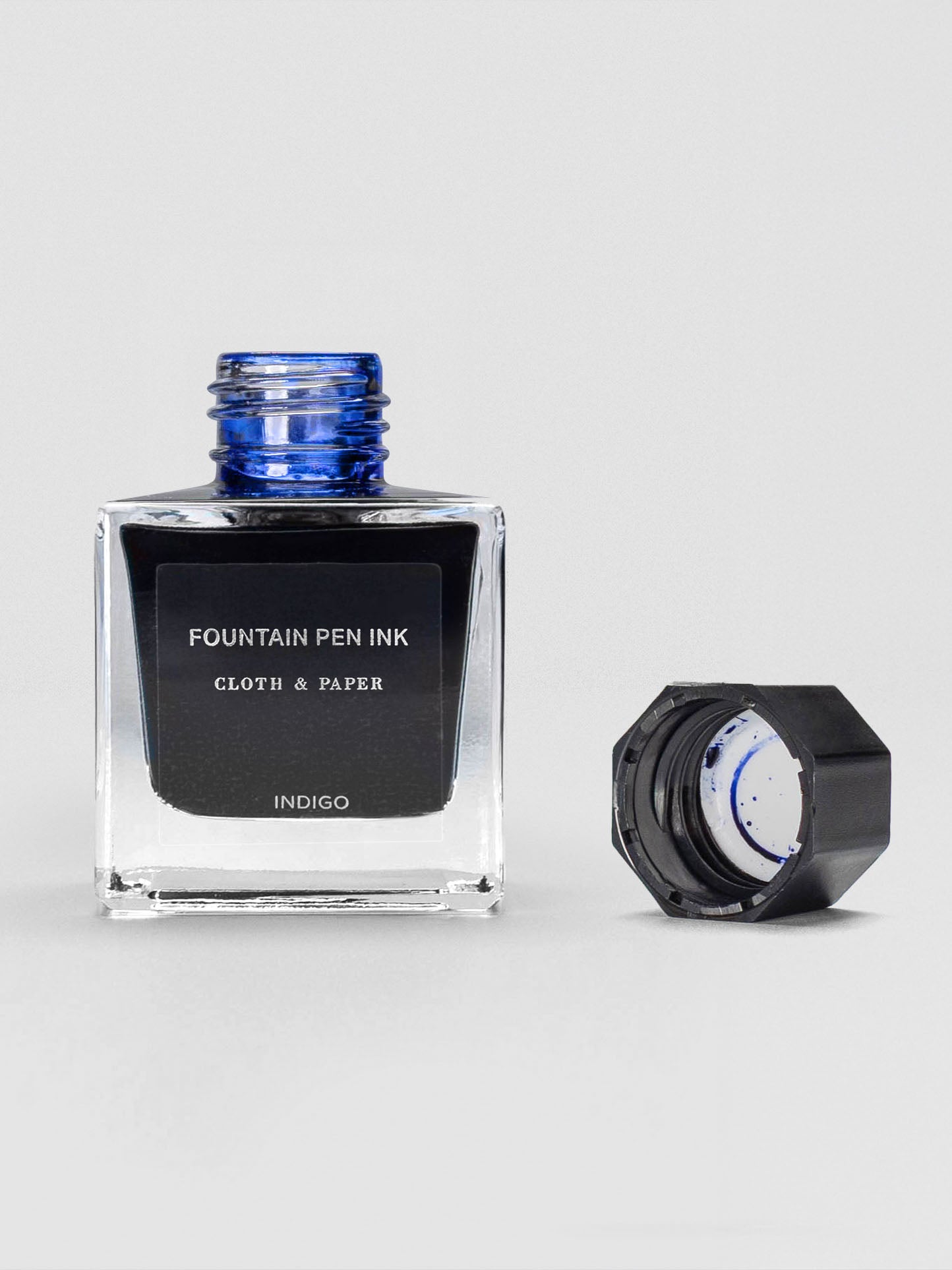 A square glass bottle of Cloth & Paper Fountain Pen Ink in Indigo with the cap removed, revealing the blue-tinted bottle opening and ink residue inside the cap.