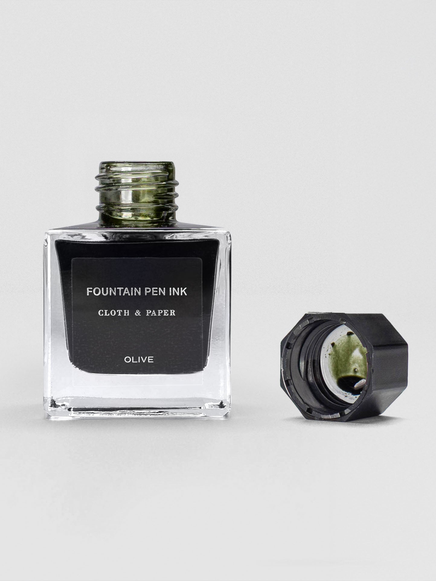 A square glass bottle of Cloth & Paper Fountain Pen Ink in Olive with the cap removed, showing a green tint around the bottle neck and ink inside the cap.