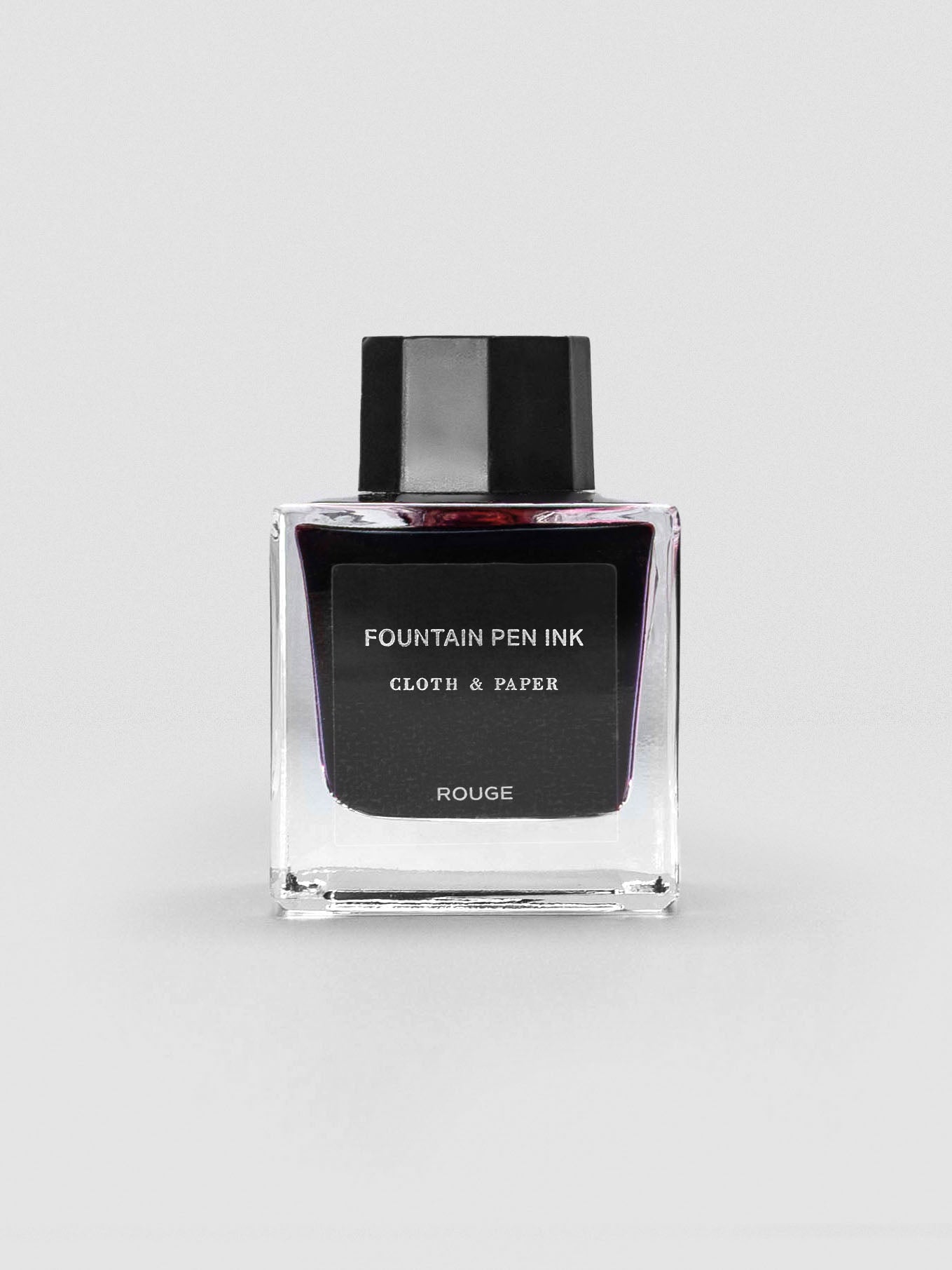 A square glass bottle of Cloth & Paper Fountain Pen Ink in Rouge, featuring a deep red tint in the clear glass and a minimalist black label with black cap.