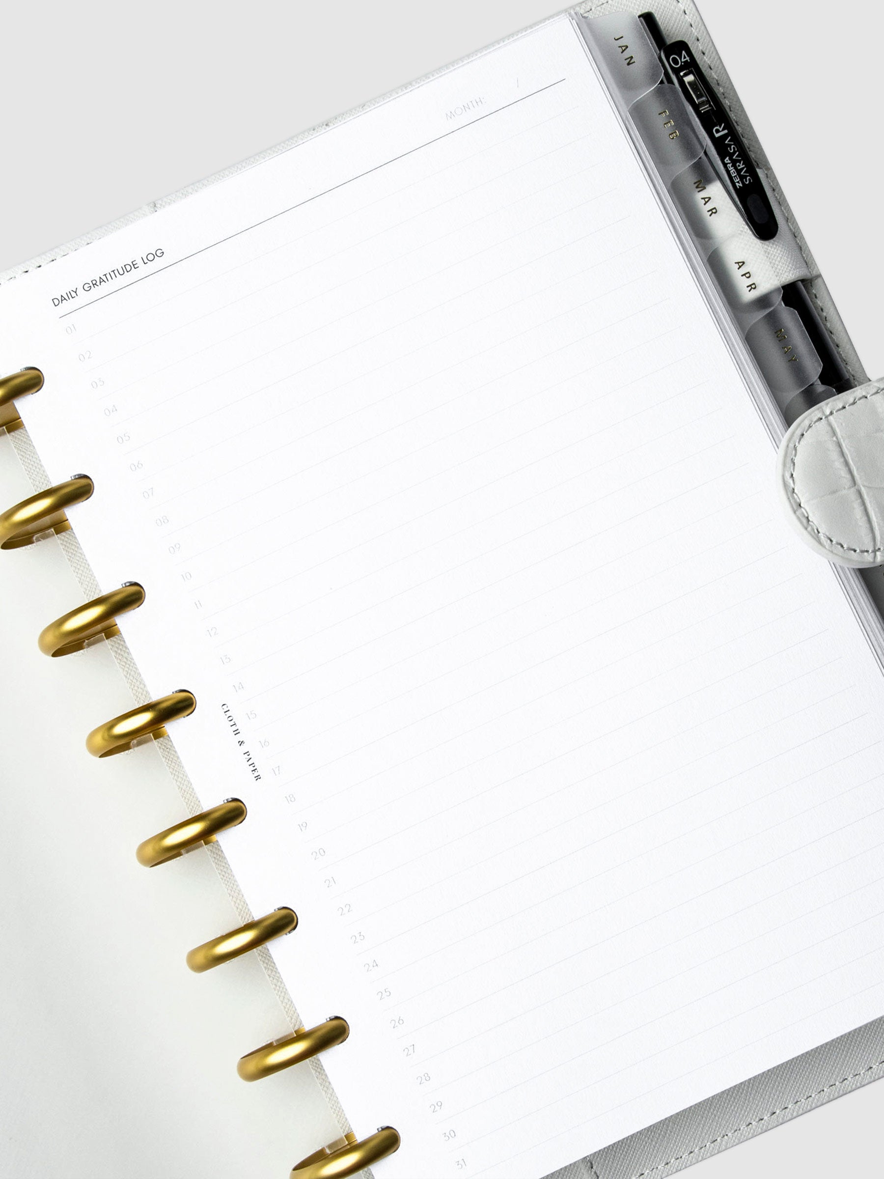Daily Gratitude Log Planner Inserts styled inside a discbound planner in a white leather agenda cover. The discbound planner has gold rings, and there is black pen tucked into the agenda cover's pen loop.