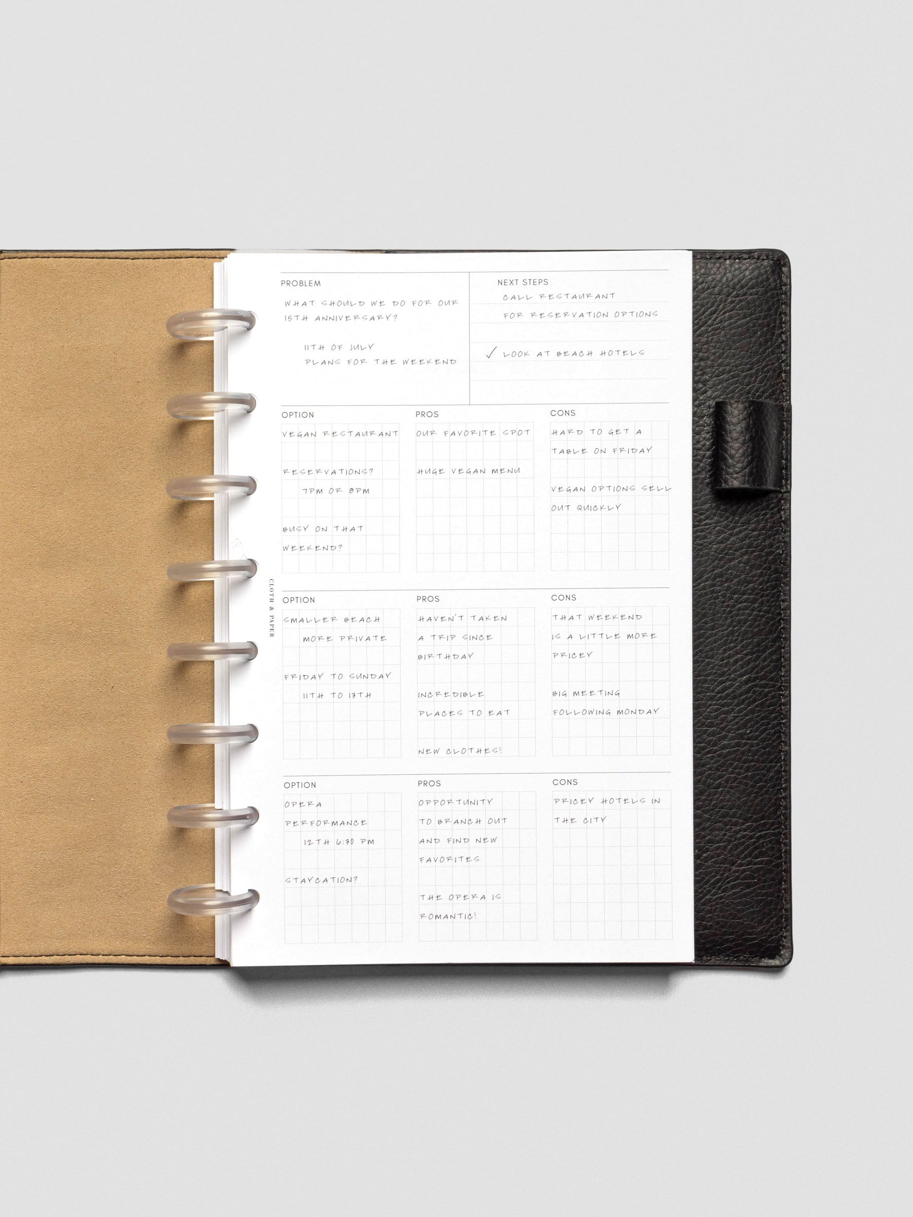 Half-letter decision-making insert inside a black leather planner with tan interior, filled with handwritten notes across problem, next steps, and option grids with pros and cons.
