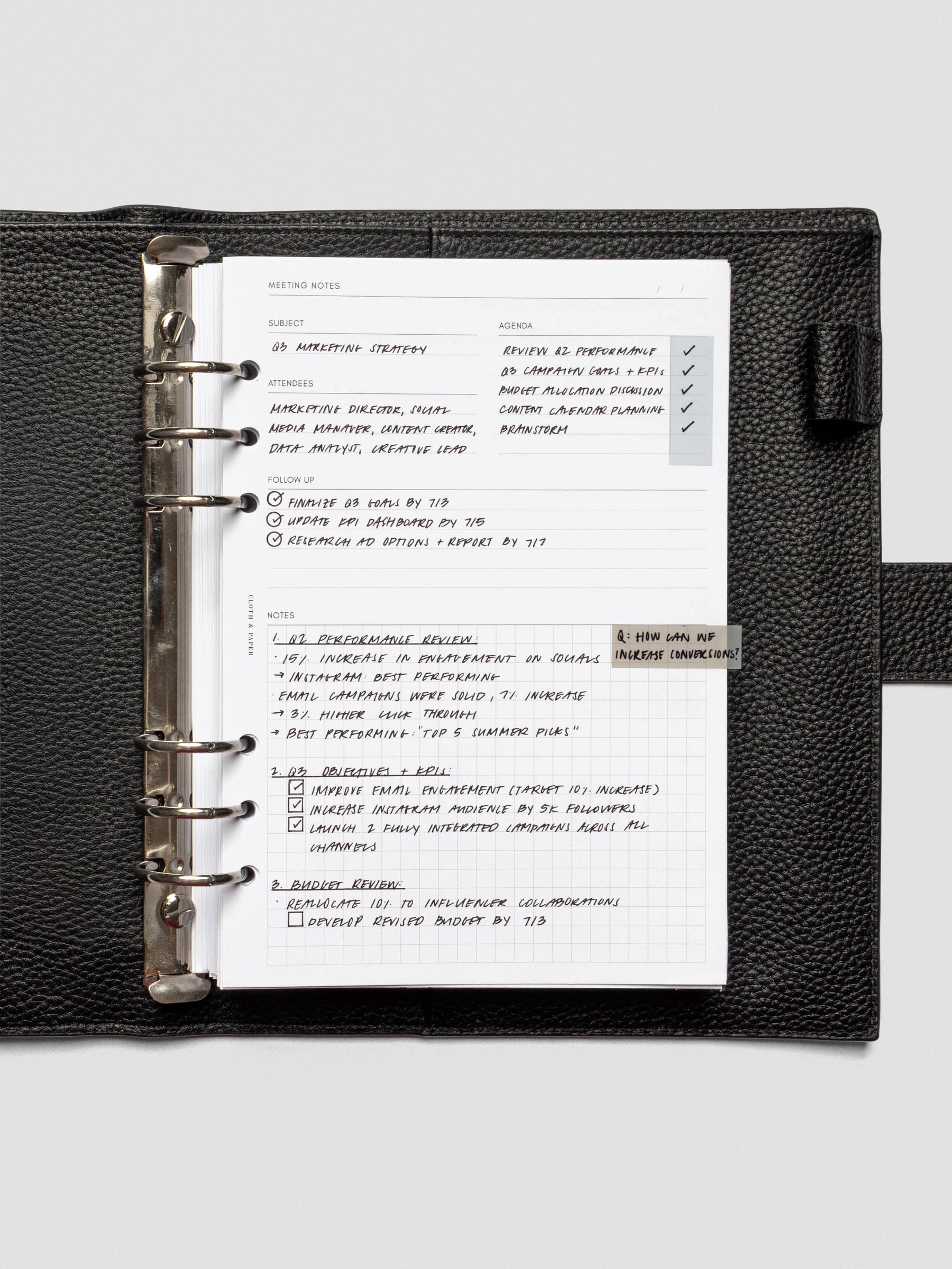 A meeting notes insert neatly filled with handwritten notes, secured in a black leather six-ring planner. The structured layout includes subject, agenda, attendees, follow-up, and a gridded notes section.