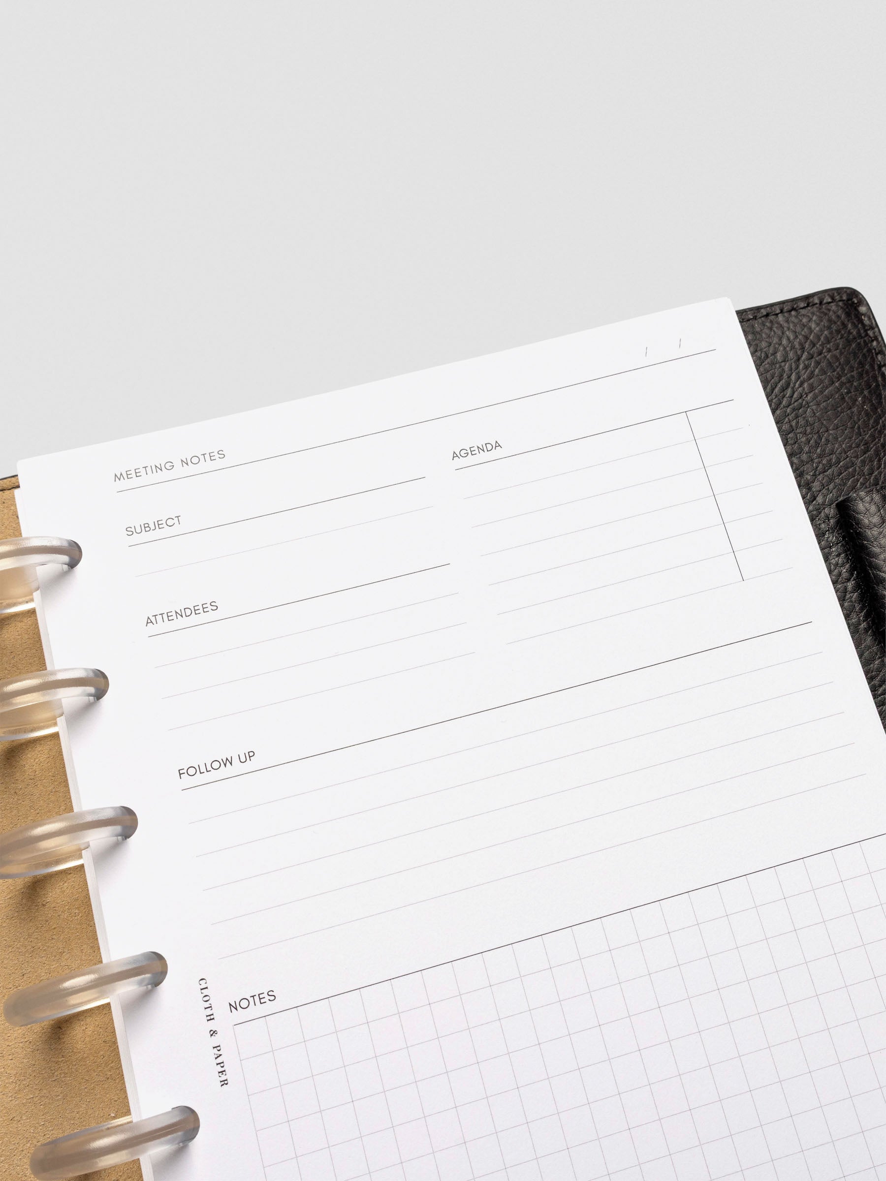 Close-up of a meeting notes insert with a minimal layout for subject, agenda, attendees, follow-up, and notes. The page is placed in a black leather planner with a tan interior and clear disc binding.