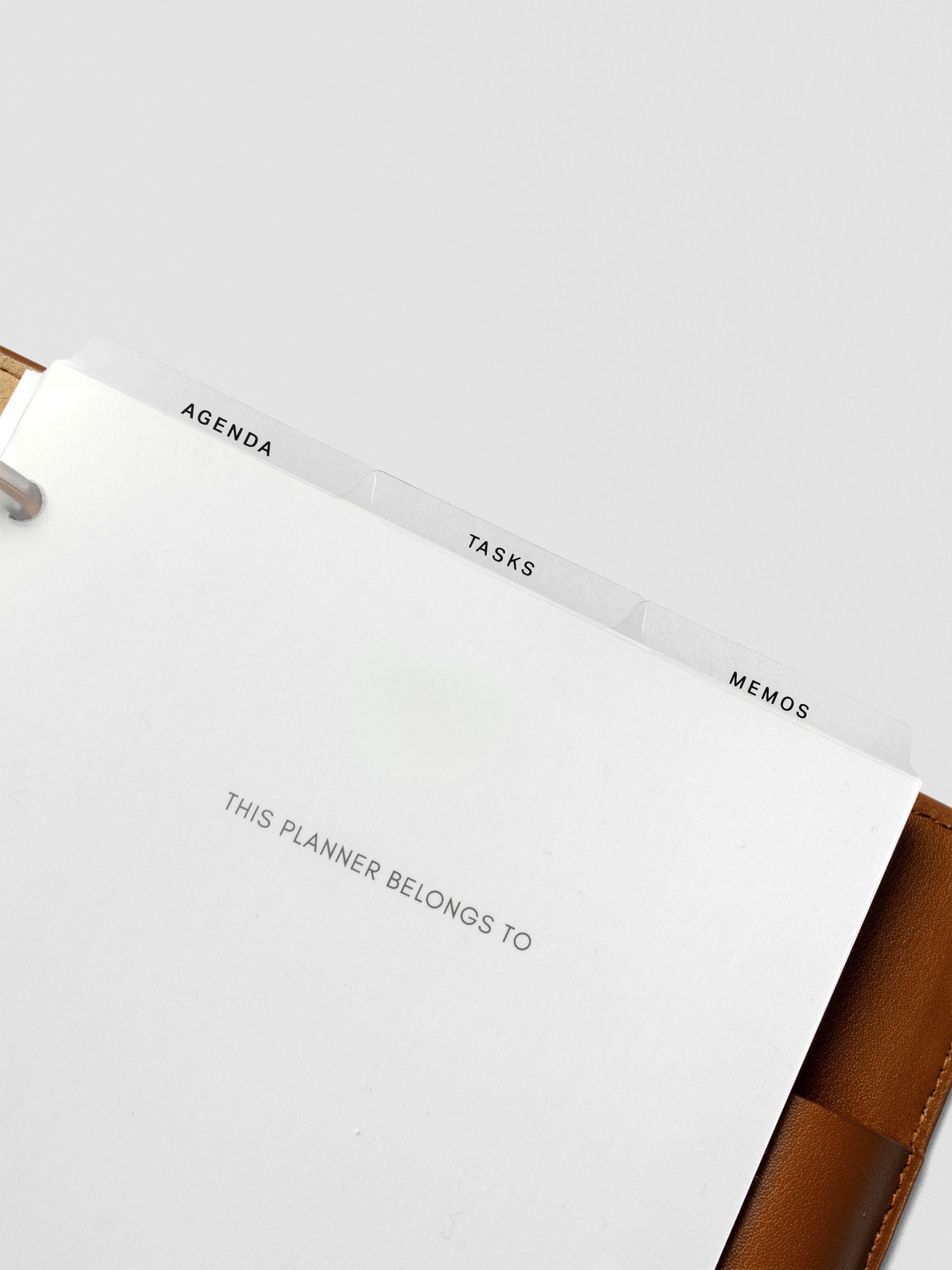 Ultra-thin clear planner dividers placed inside a tan leather planner, tabs labeled Agenda, Tasks, and Memos visible above the title page.
