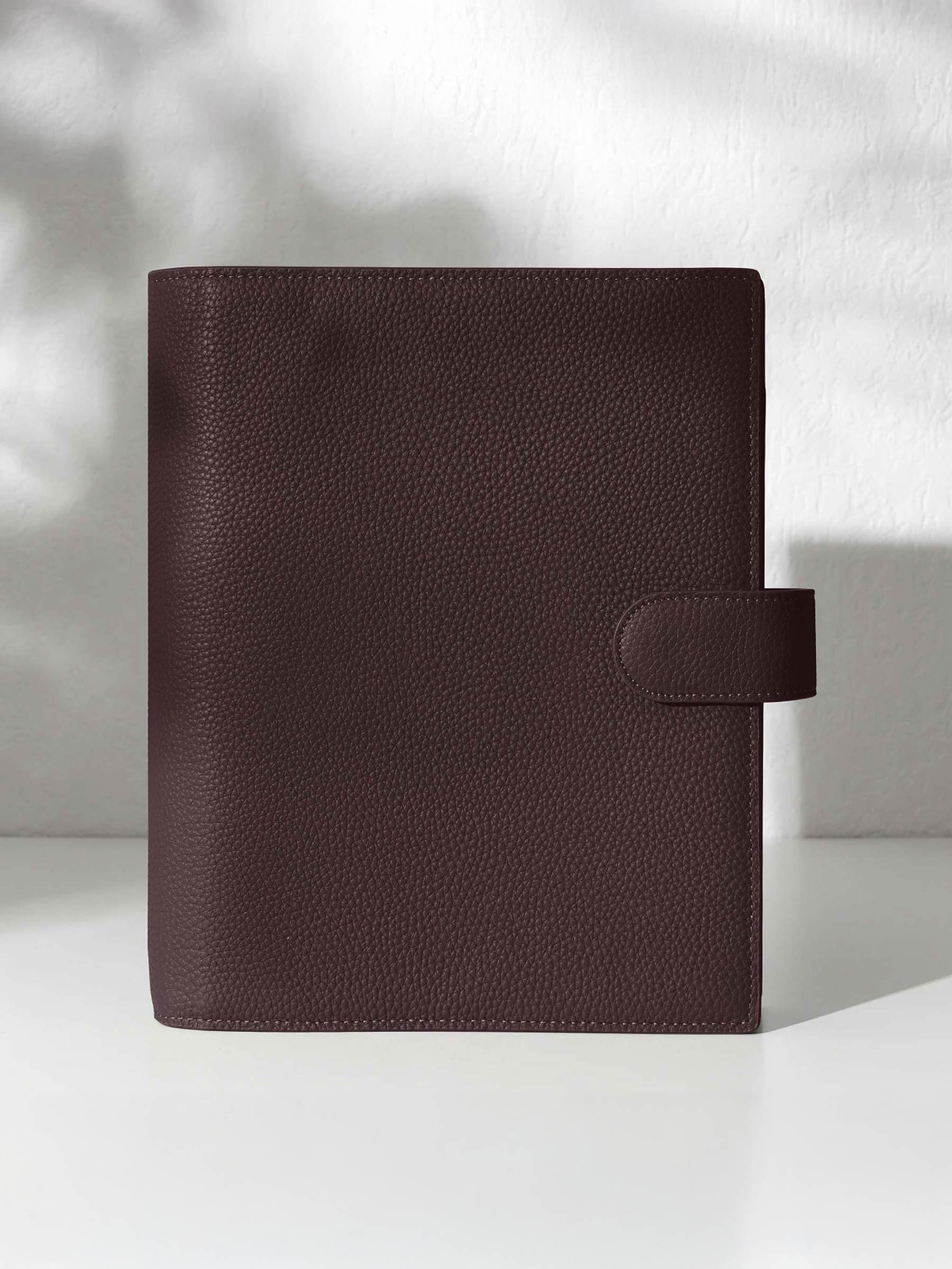 A closed dark burgundy leather planner with a closure displayed against a shadowed background.