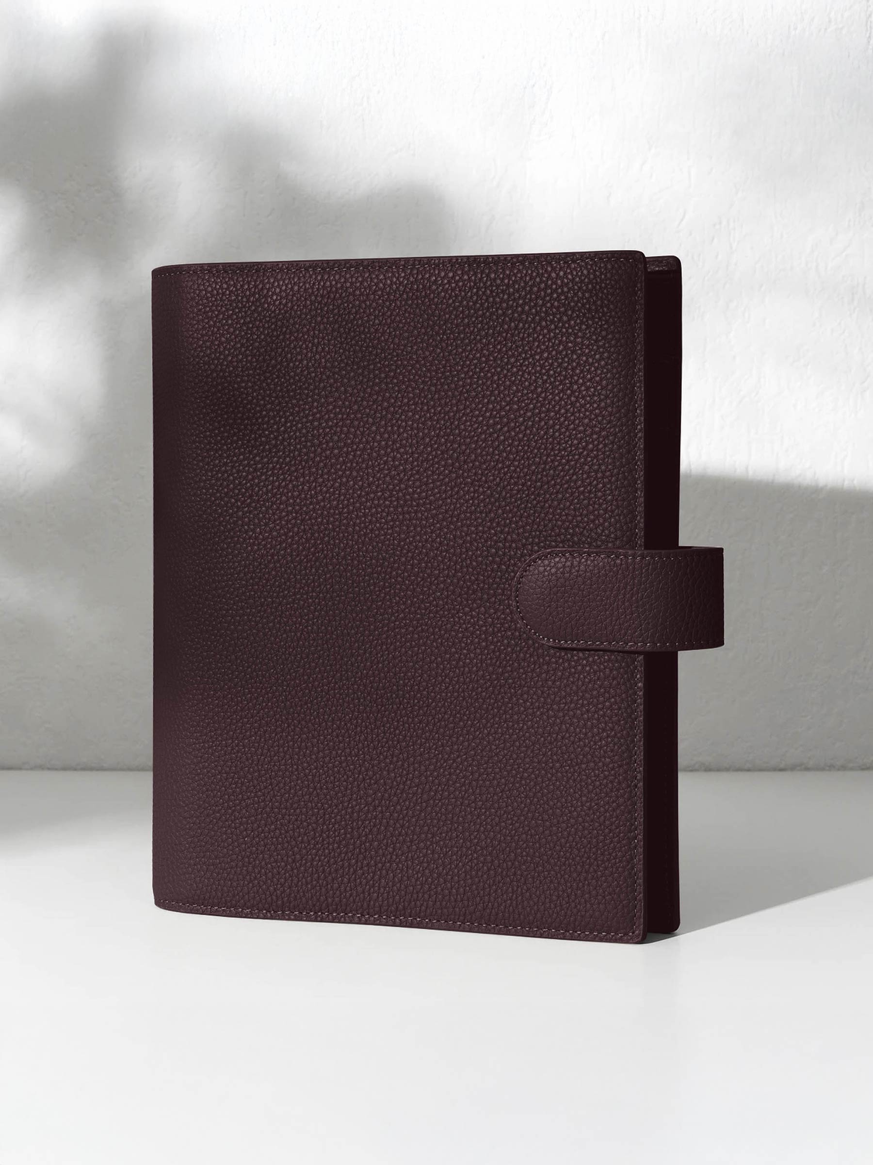 A closed dark burgundy leather planner with a closure displayed against a shadowed background.