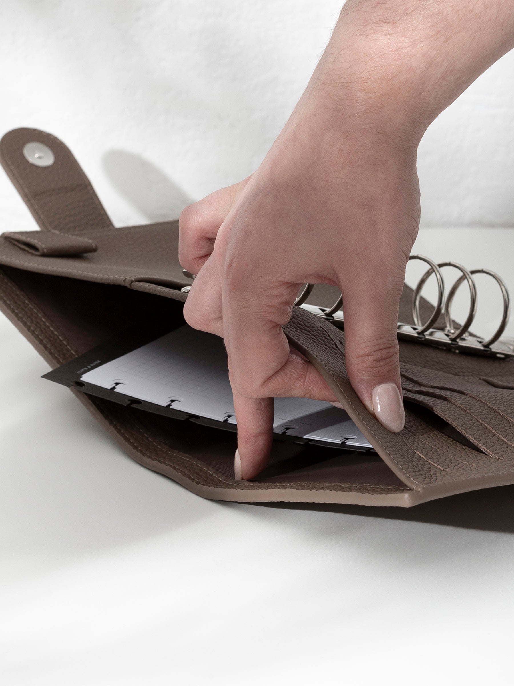 A fair-skinned hand shown opening the back pocket of the brown leather agenda on an off-white background. There are inserts and a dashboard inside the pocket.