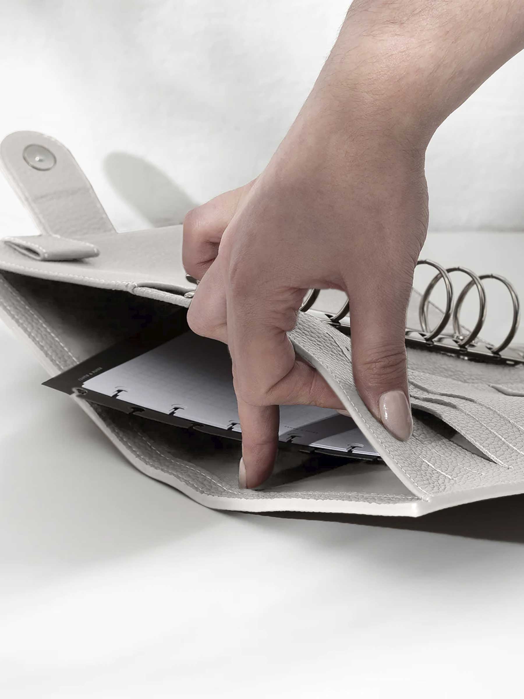 A fair-skinned hand shown opening the back pocket of the ash leather agenda on a white background. There are inserts and a dashboard inside the pocket.