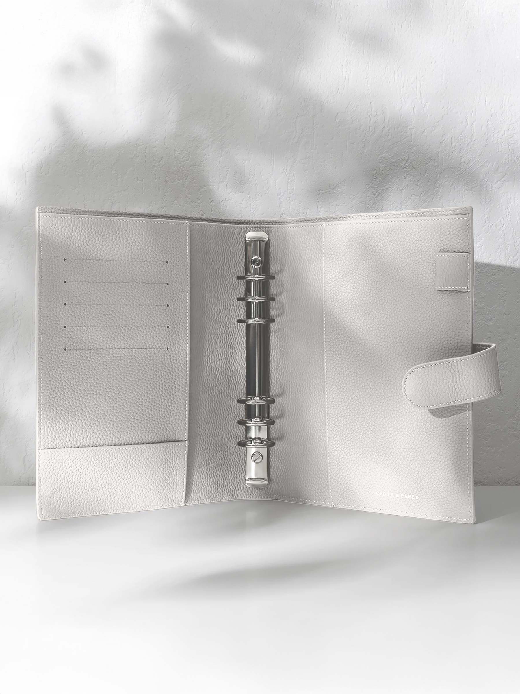 Ash leather agenda displayed on an off-white background. It is opened to display the silver hardware and company foil stamp.