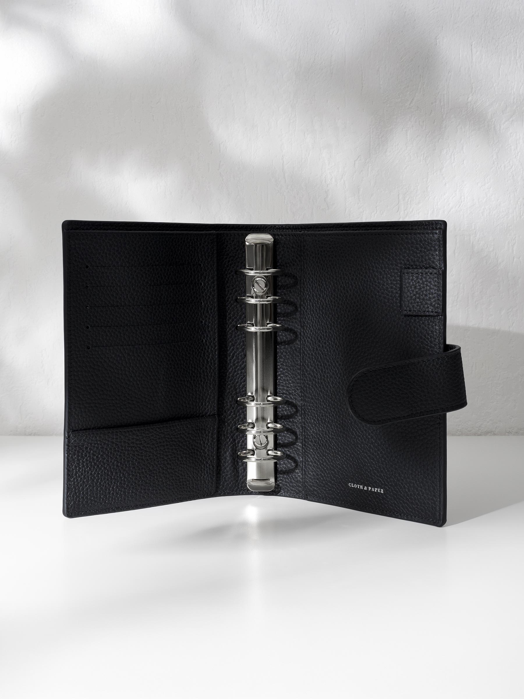 Black leather agenda displayed against a shadowed background. It is opened to show off the silver hardware and company logo stamp.